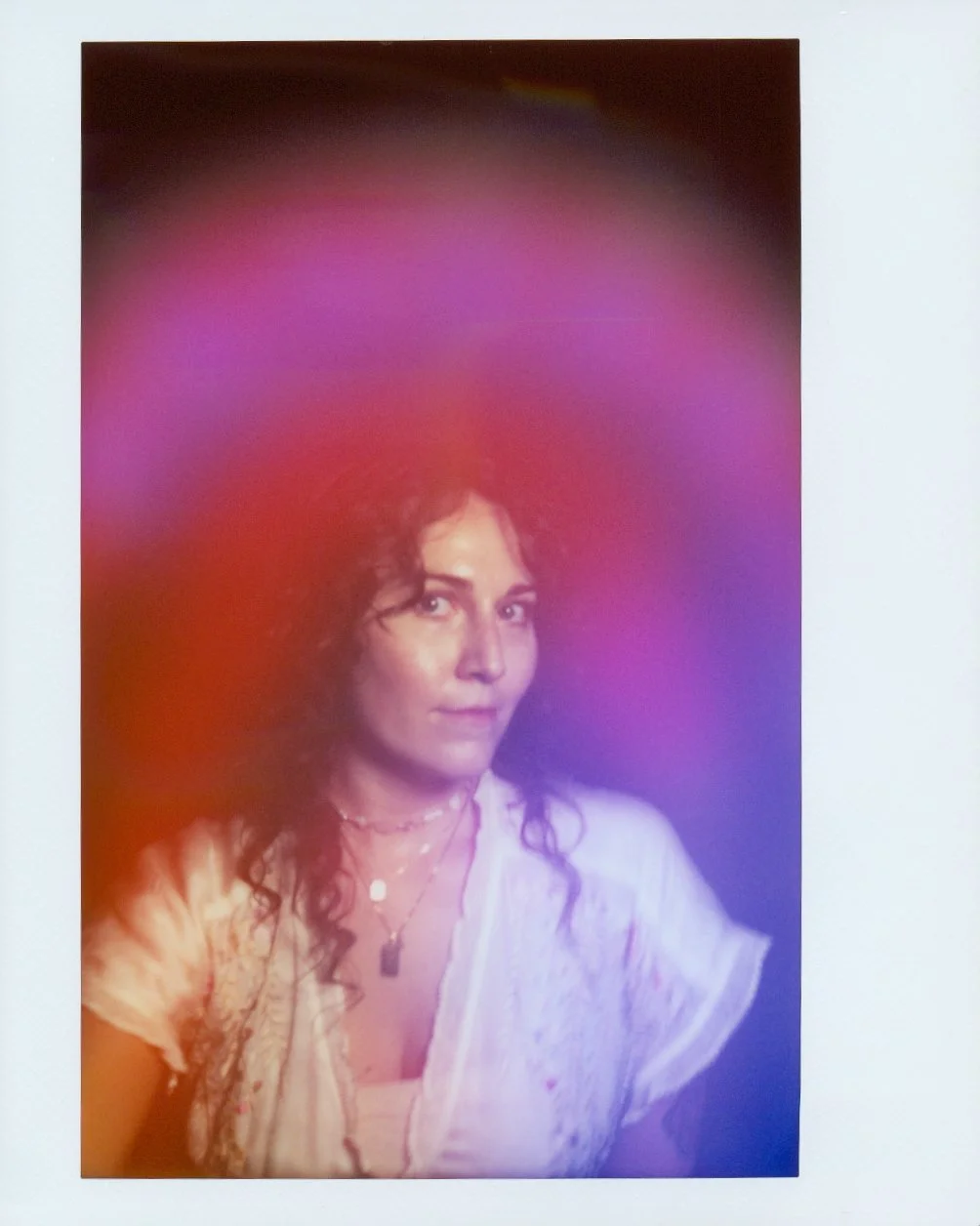 Portrait of a woman with dark, curly hair wearing a white top and layered necklaces, with a colorful purple and red light effect in the background.