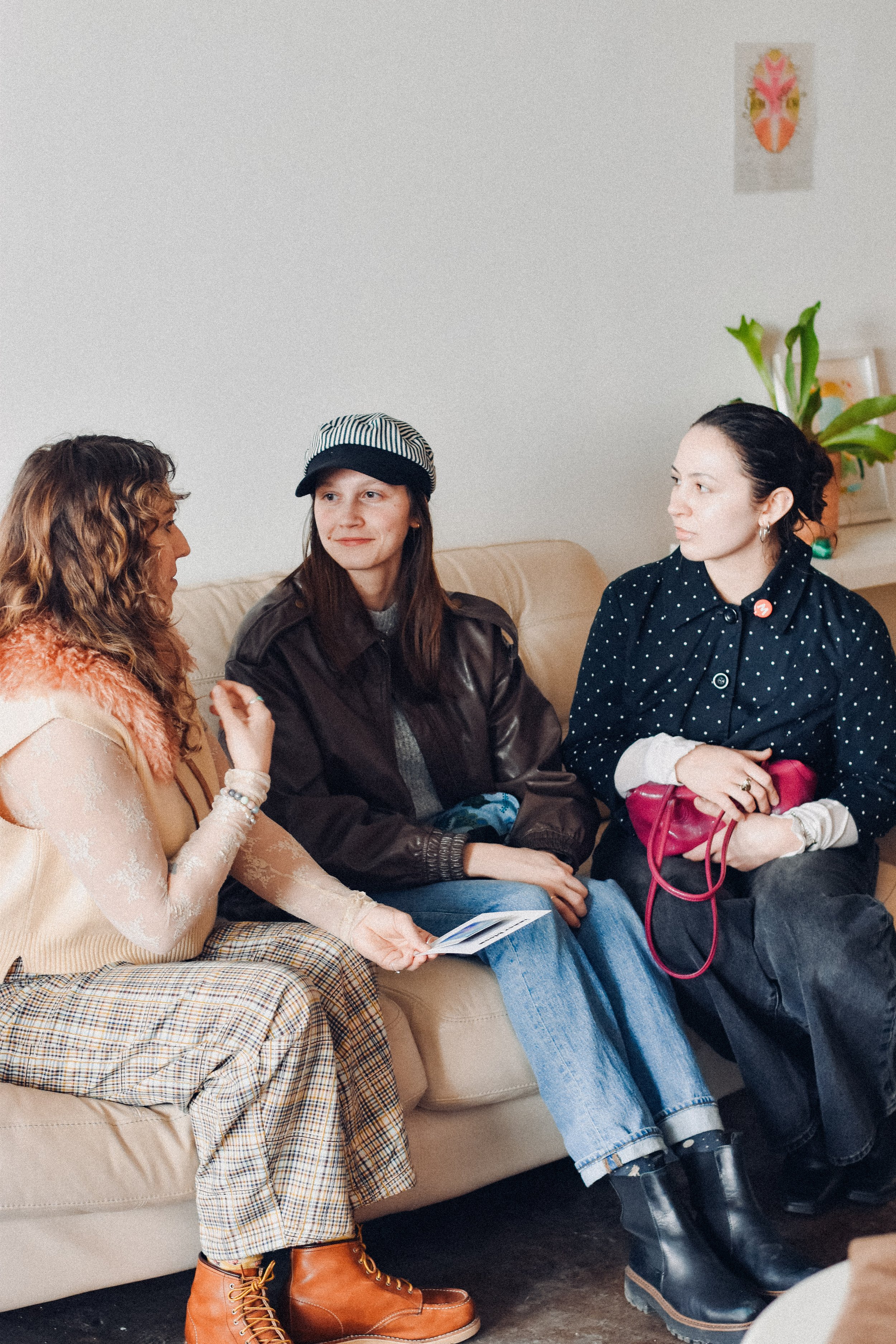 Three women sit on a beige couch in conversation inside a room decorated with artwork and plants.