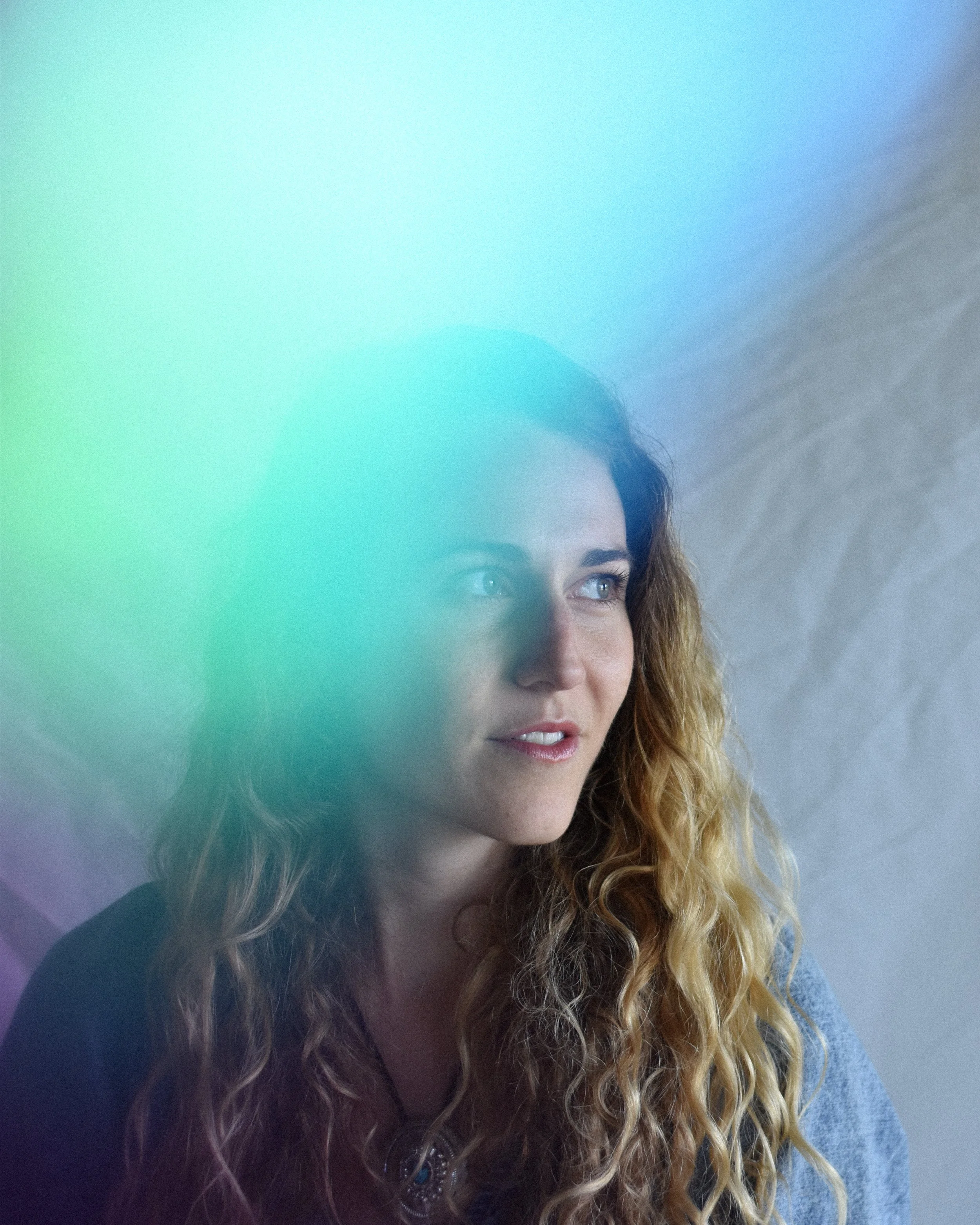 A woman with long curly blonde hair, looking to the right, with a soft focused, colorful overlay in the foreground.
