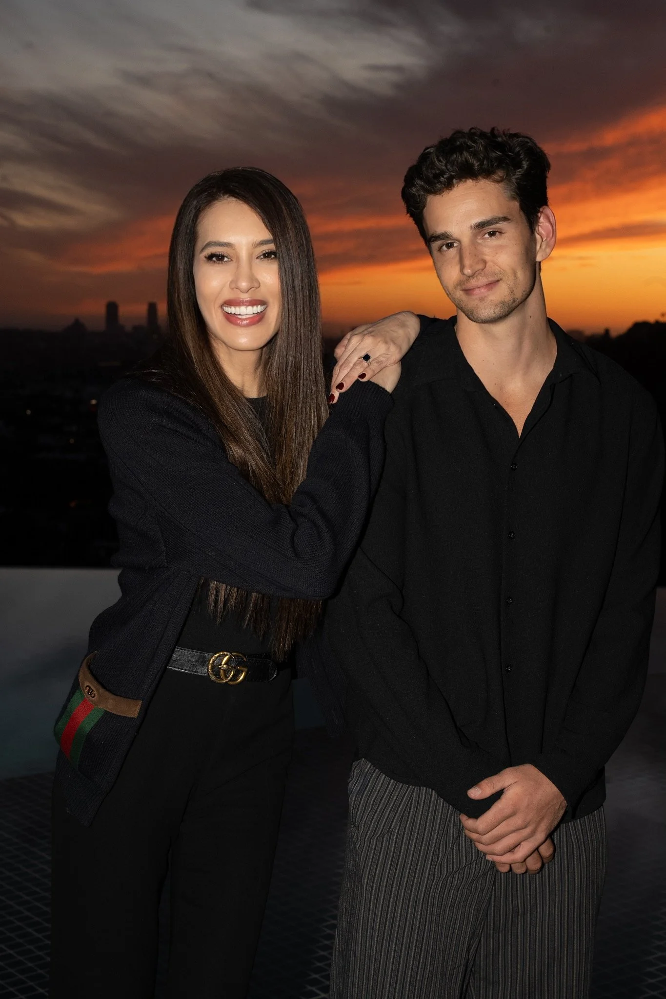 A smiling woman with long brown hair and a man with curly hair pose together during sunset, with a city skyline in the background.