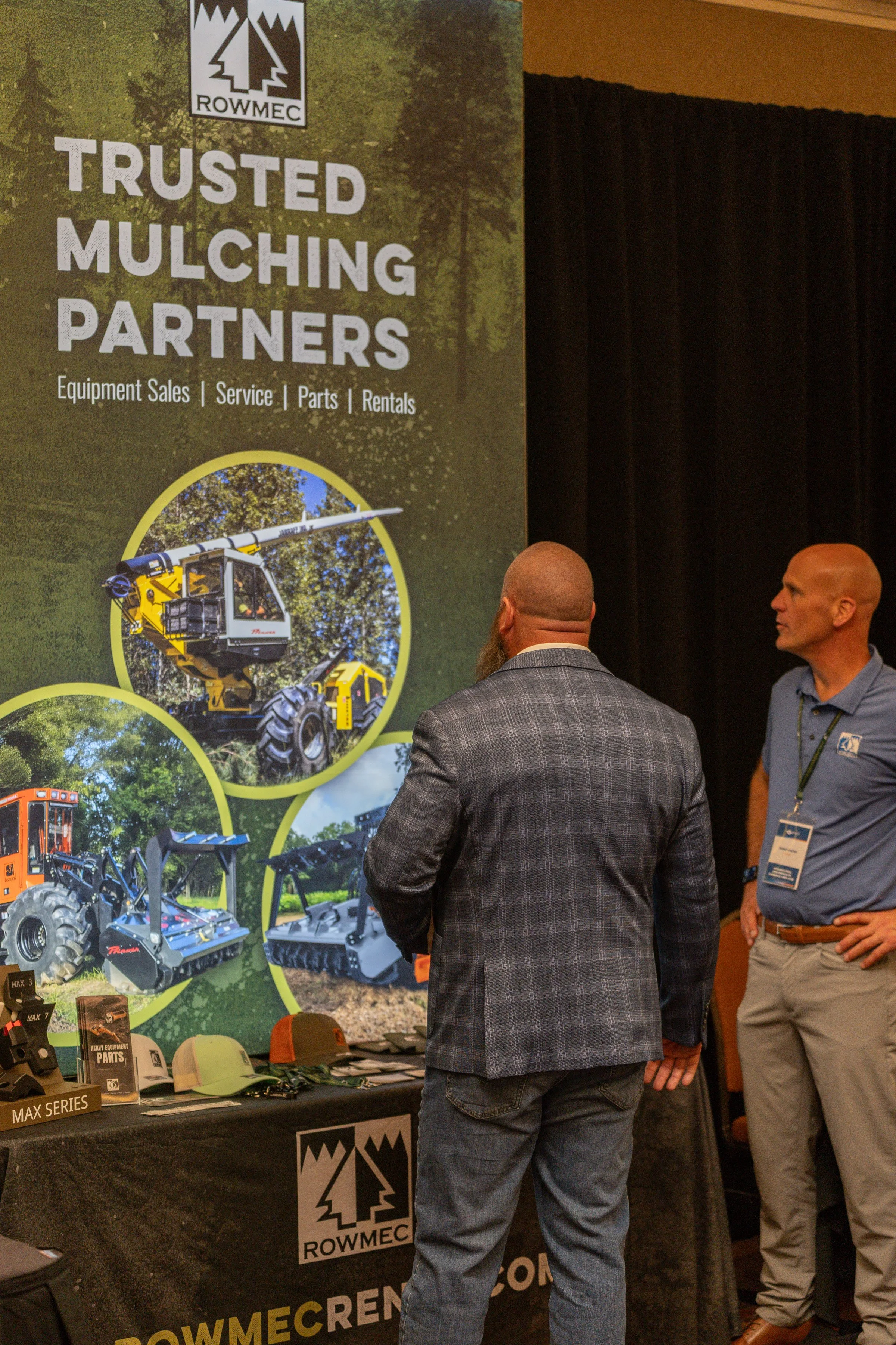 A promotional display for ROWMEC, showcasing equipment and parts for mulching including a large sign that reads 'Trusted Mulching Partners,' with images of various machinery such as a cherry picker, an excavator, and other equipment, with two men sta