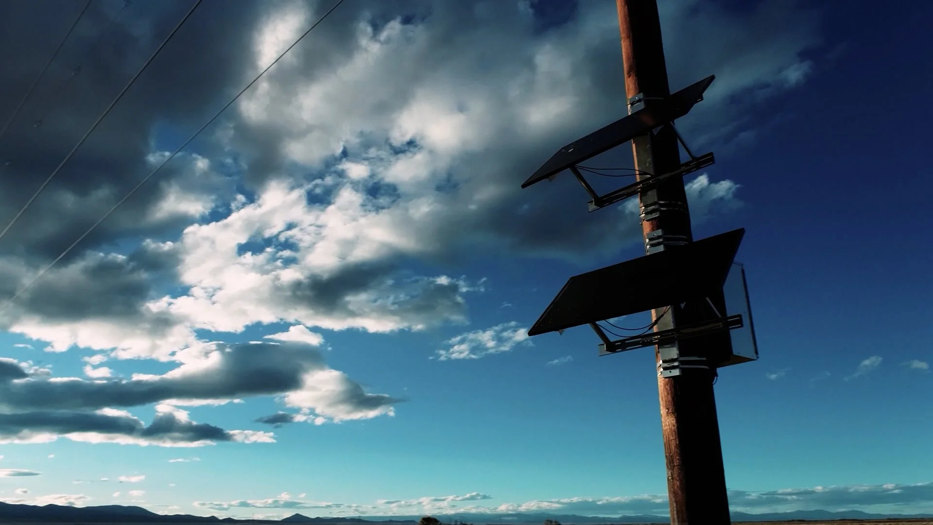A utility pole with two solar panels against a partly cloudy sky with mountains in the distance.