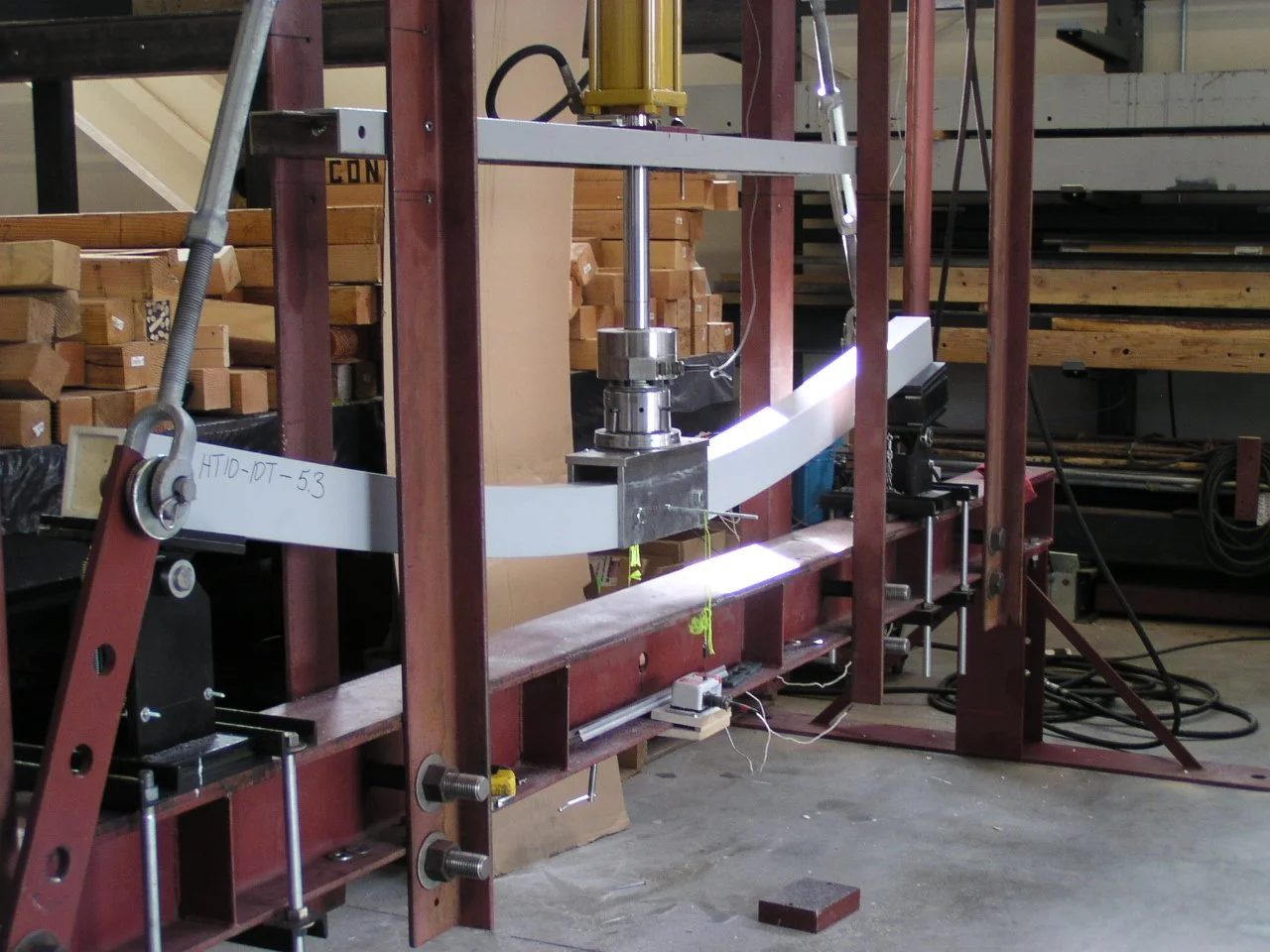 A laboratory or workshop setup with a large curved metal strip mounted on a red metal frame, connected to various sensors and wires, with wooden shelves and stacks of wood in the background.