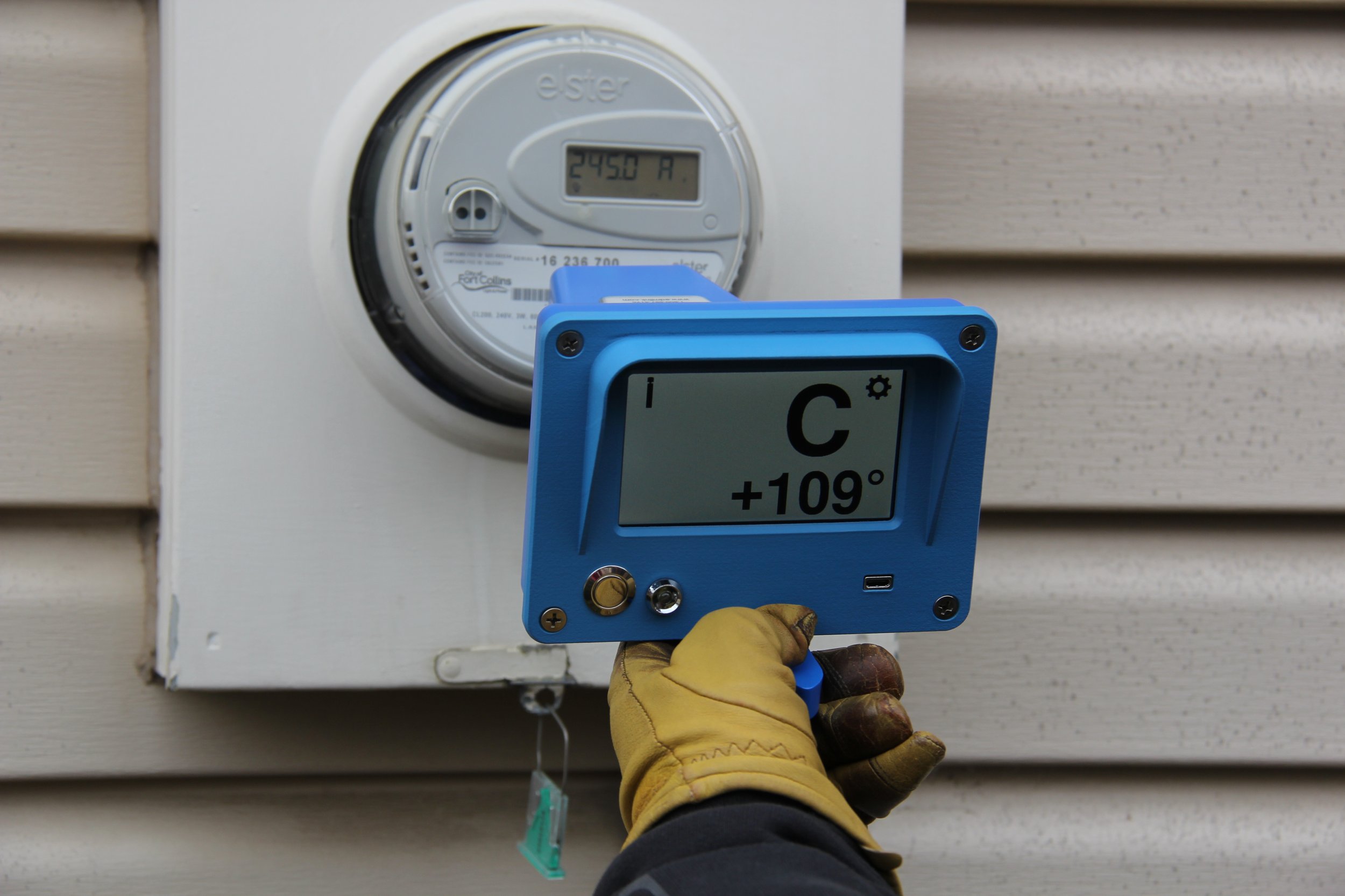 Worker wearing yellow gloves holding a blue digital refrigeration or air conditioning service tool outside a house, with a digital thermostat displaying +109°F on the wall.