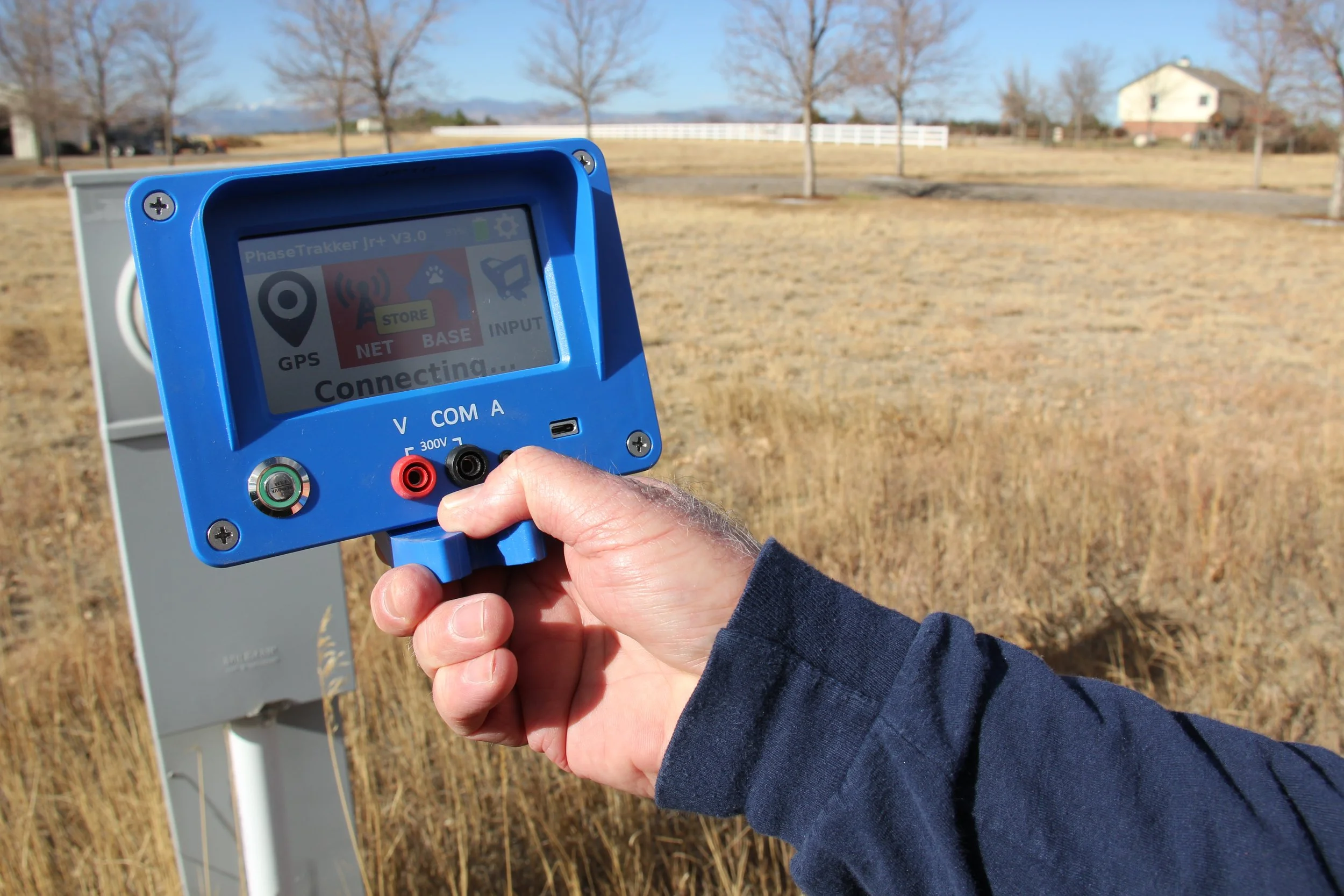 Person holding a blue GPS device outdoors in a grassy field with trees and a house in the background.