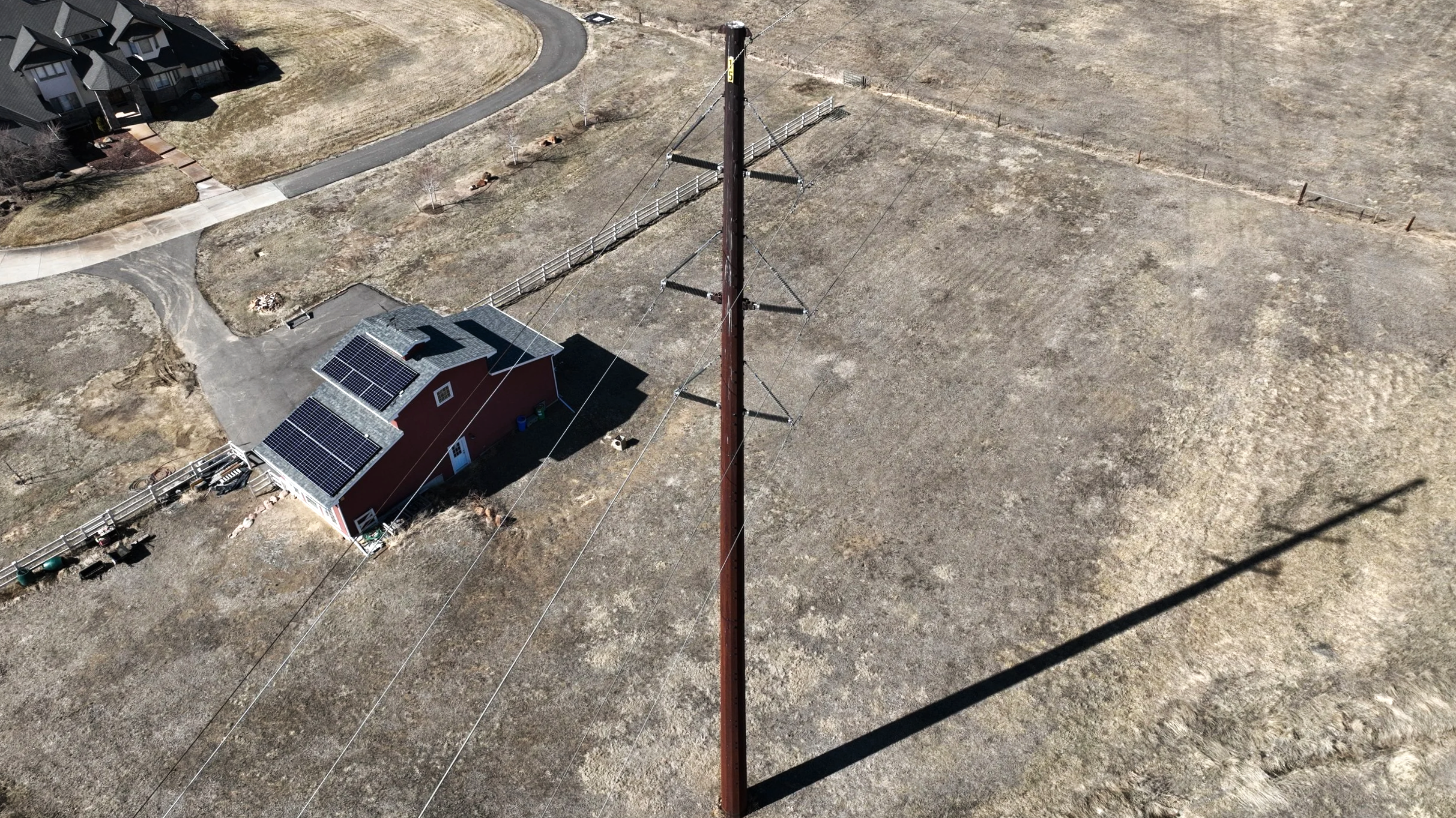 A red house with solar panels on the roof, a utility pole with power lines, and a large vacant lot with patches of grass and dirt.