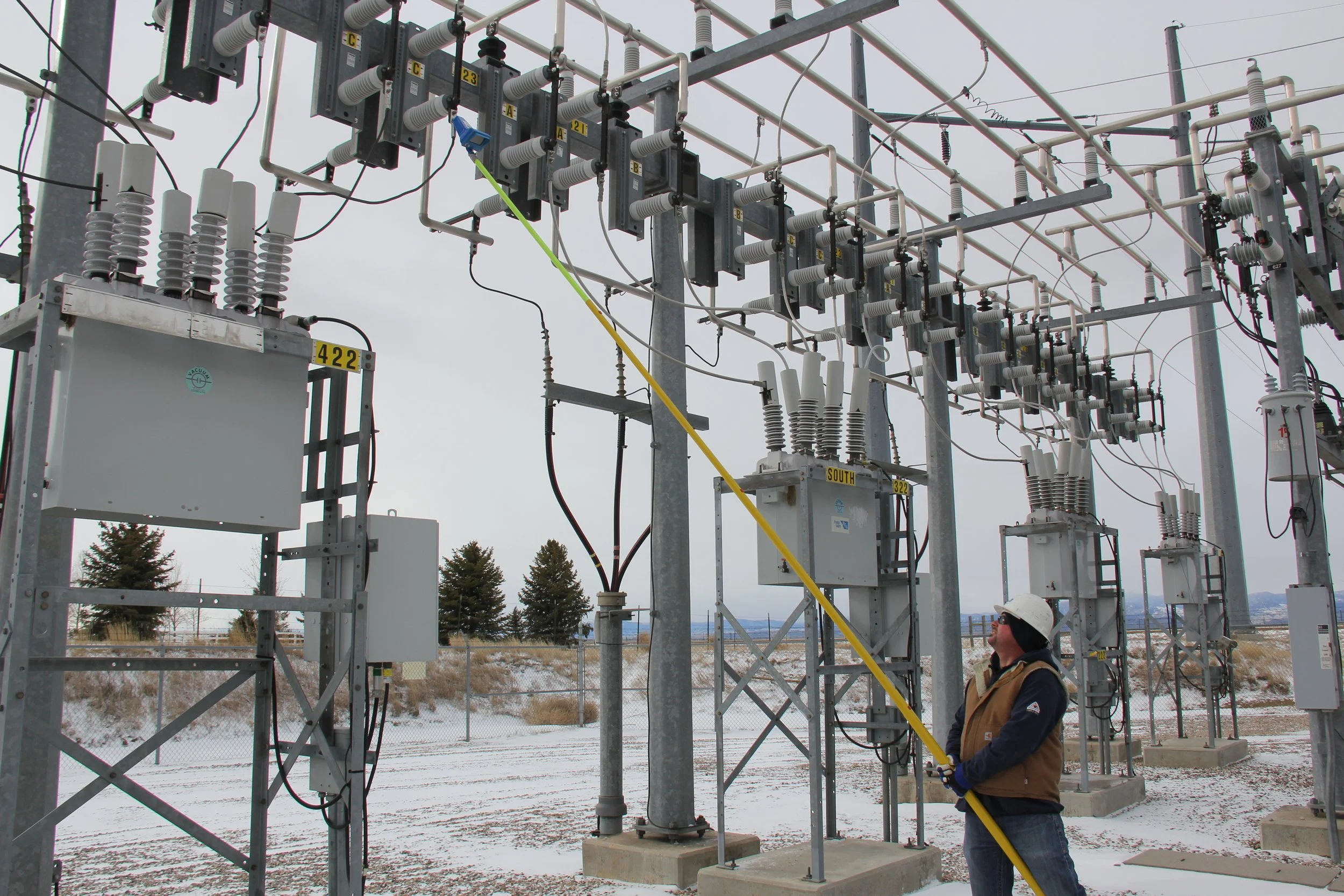 A worker wearing a hard hat and safety vest inspecting electrical equipment at an outdoor power substation surrounded by metal framework and electrical insulators under cloudy skies.