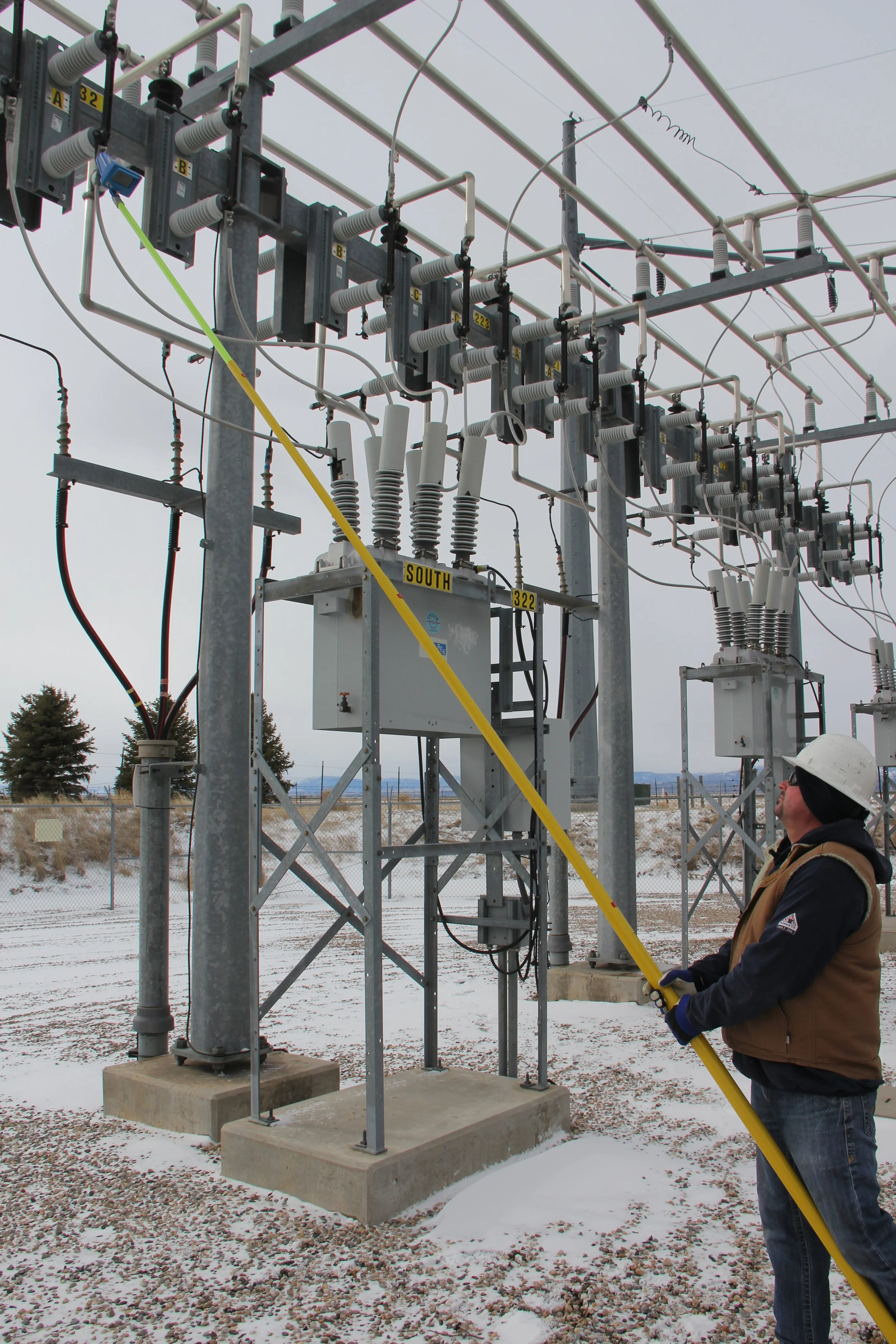 A man wearing a hard hat and brown jacket working on electrical equipment outdoors in snowy weather, using a long yellow tool.