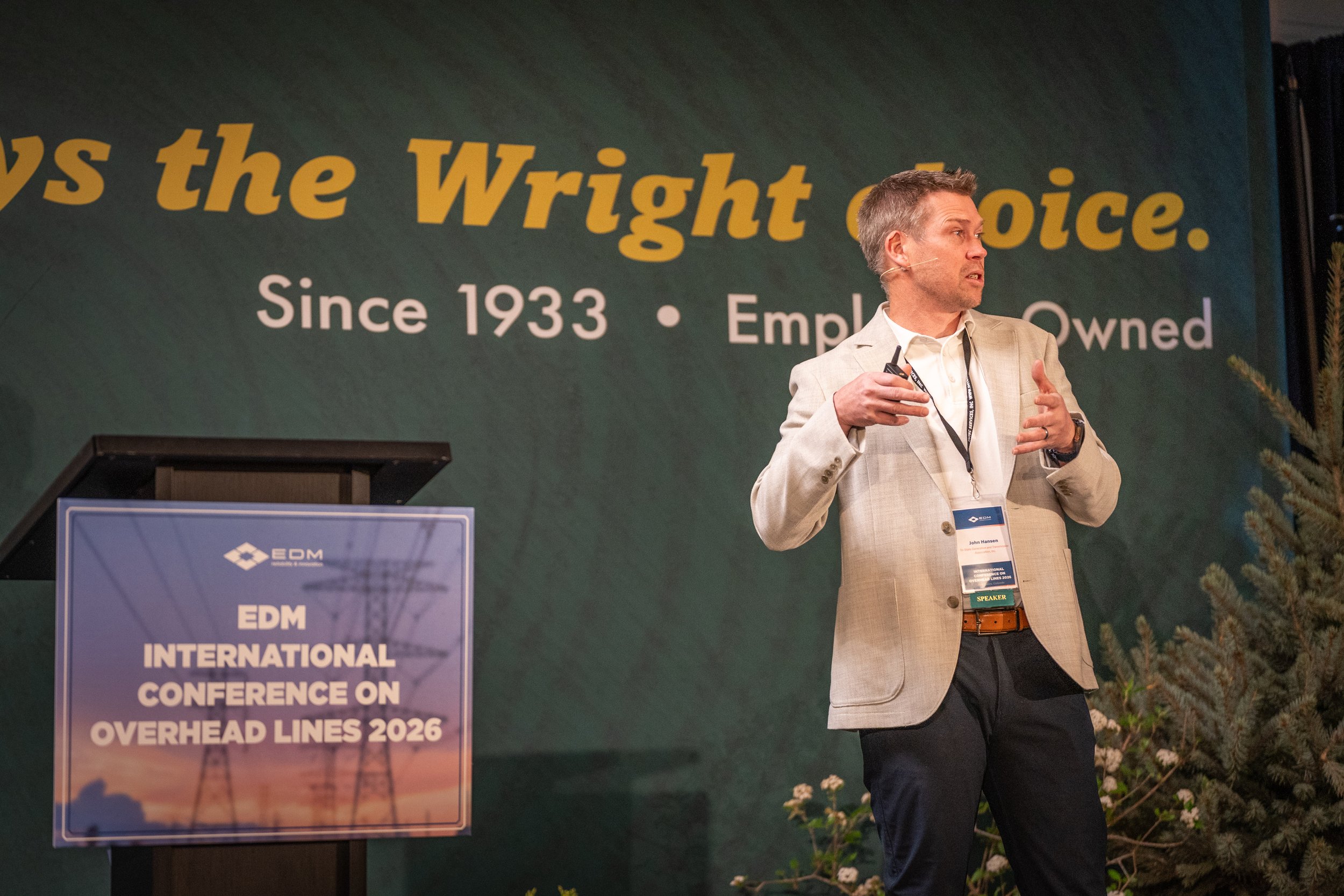 A man giving a presentation on stage at an international conference, with a large sign behind him that reads 'Since 1933 - Employee-Owned'. The man is wearing a light-colored blazer and has a headset microphone.
