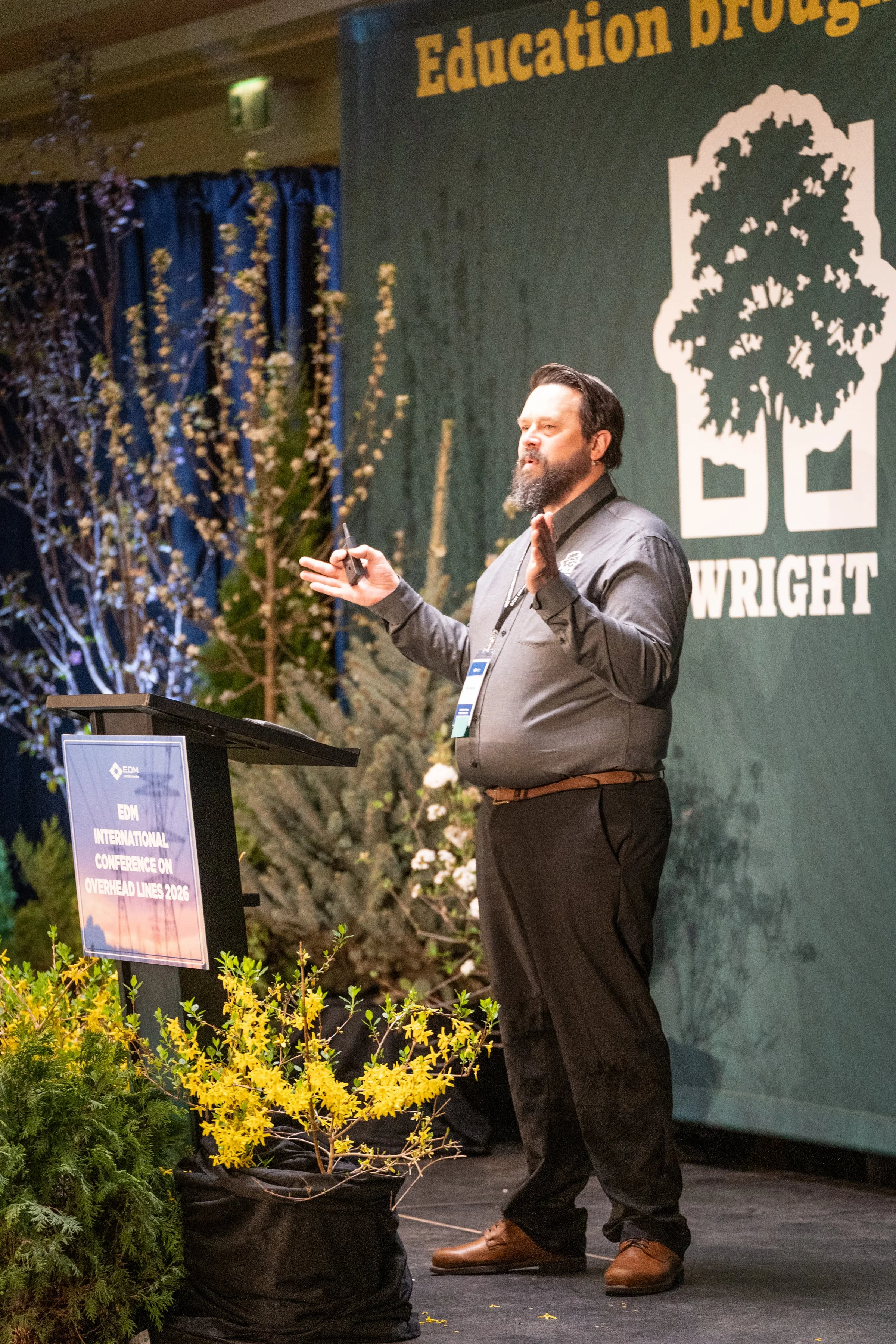 A man with a beard and dark hair, wearing a dark shirt and black pants, is giving a presentation on a stage at the EDM International Conference on Overhead Lines 2026. He is making a hand gesture with one hand and holding a clicker in the other. Behi