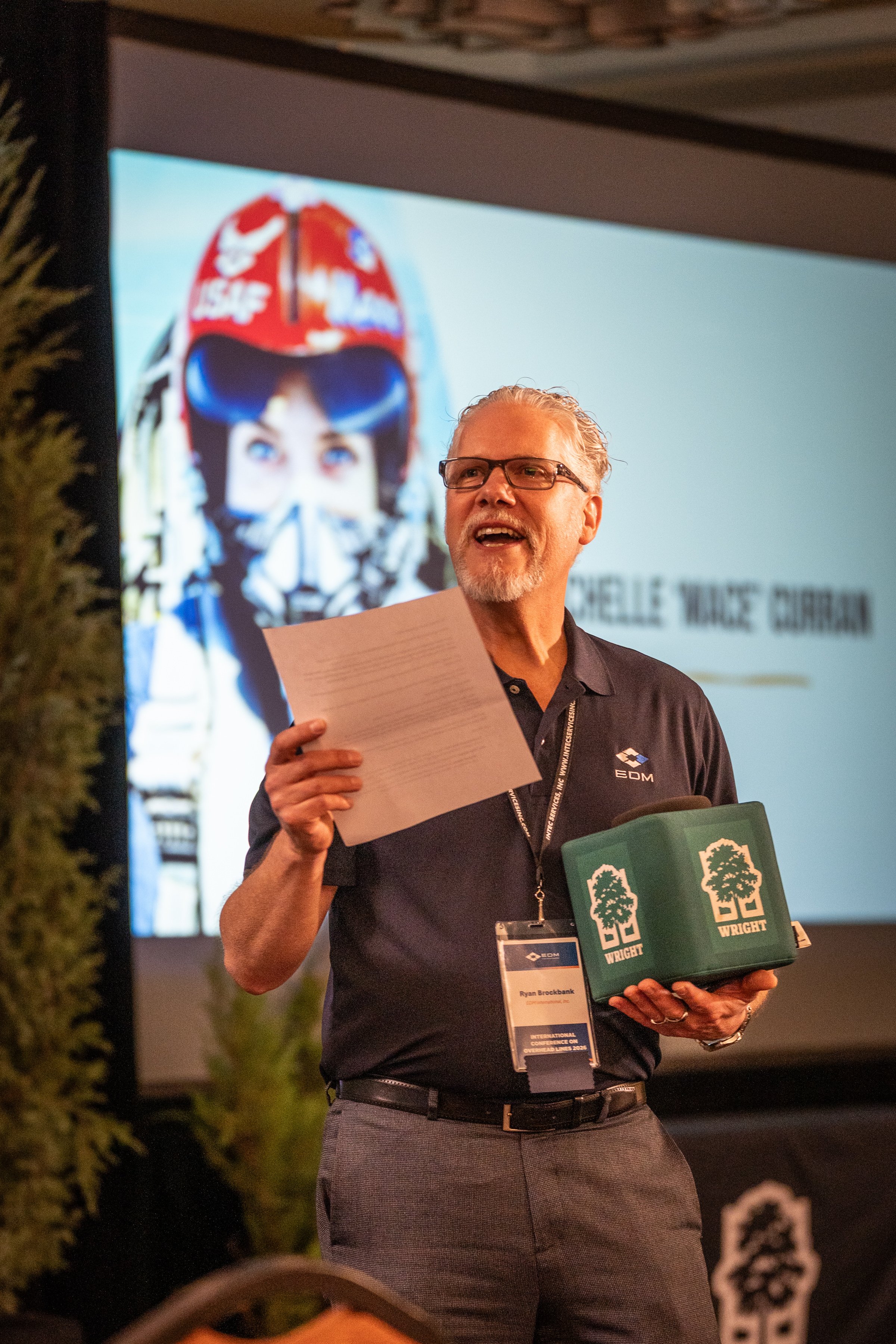A man with glasses holding a piece of paper and a green box with trees and the word 'WRIGHT' on it, standing in front of a screen displaying a woman in a racing helmet. The man is wearing a black shirt with a logo and a conference badge.