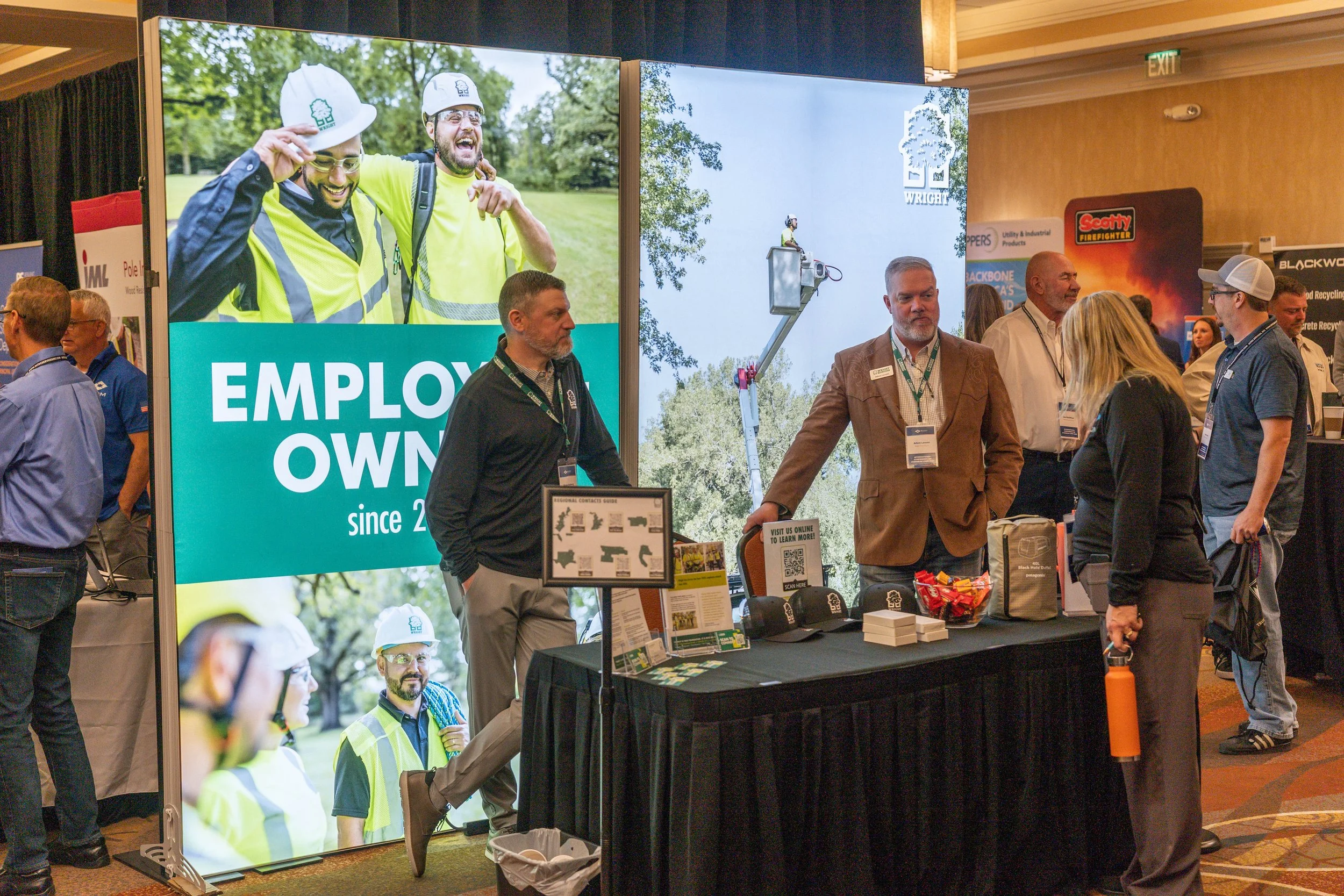 A trade show booth displaying images of workers in safety gear, including helmets and high-visibility clothing, with people engaging in conversations and browsing materials.