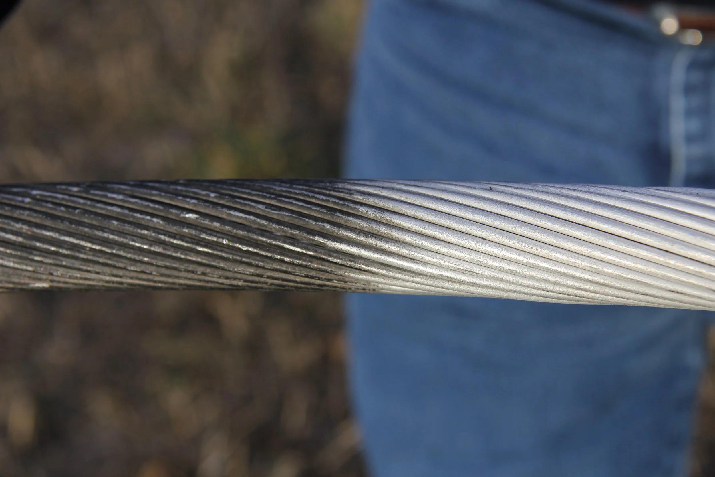 Close-up of a textured metal rod with diagonal grooves, with a blurred person wearing blue jeans in the background.