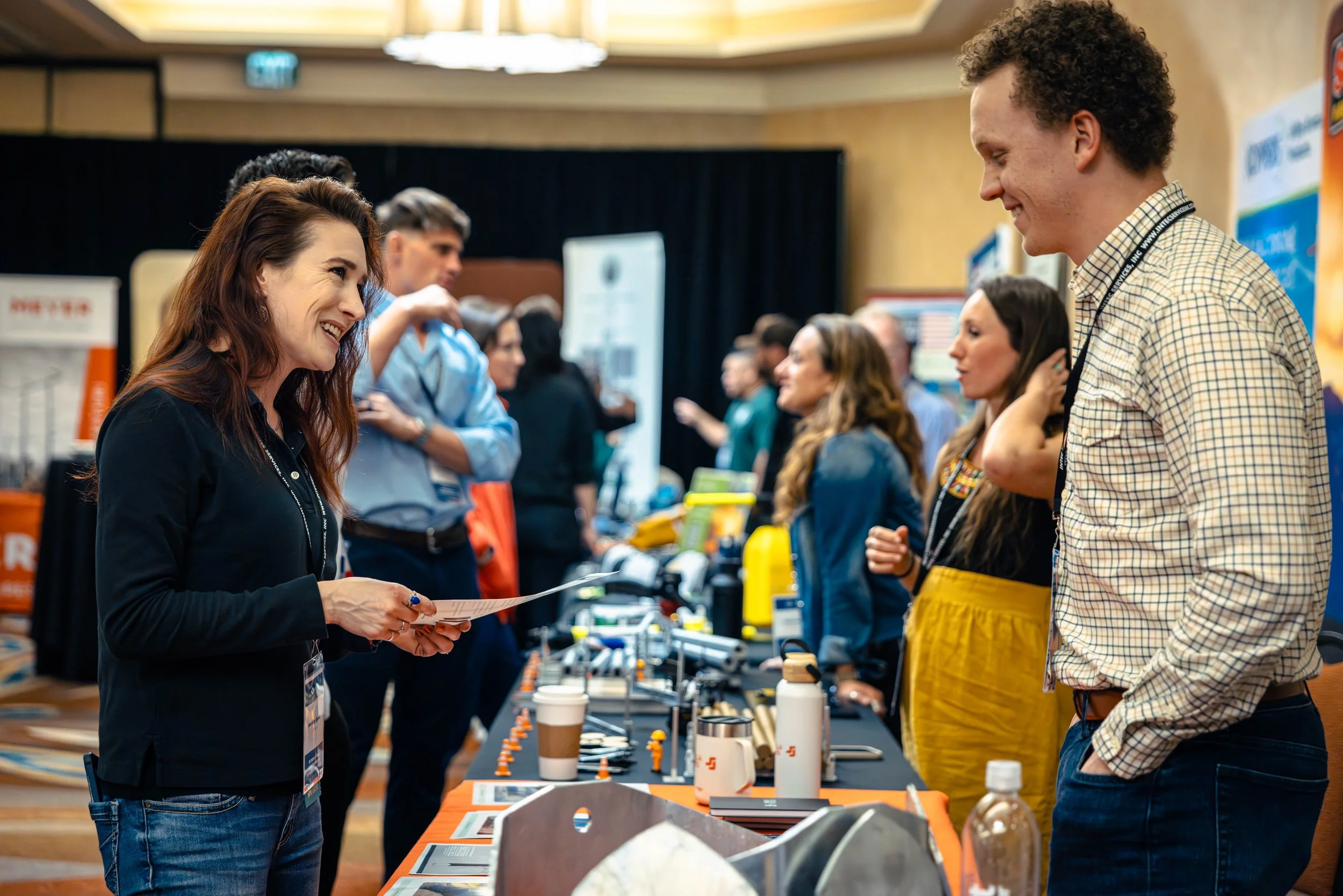 A woman with long brown hair smiling and talking to a man with curly hair at a booth in a conference or expo setting, with multiple people and display tables in the background.
