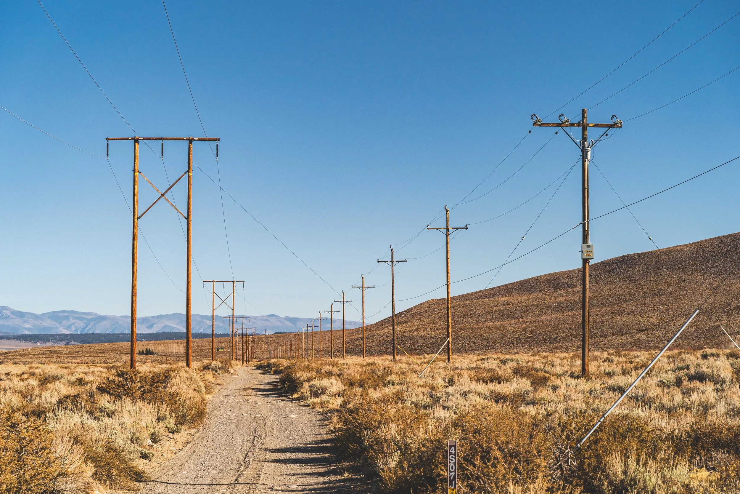A rural dirt road flanked by dry grass and sparse bushes, with wooden utility poles and power lines stretching into the distance under a clear blue sky, with mountains in the background.