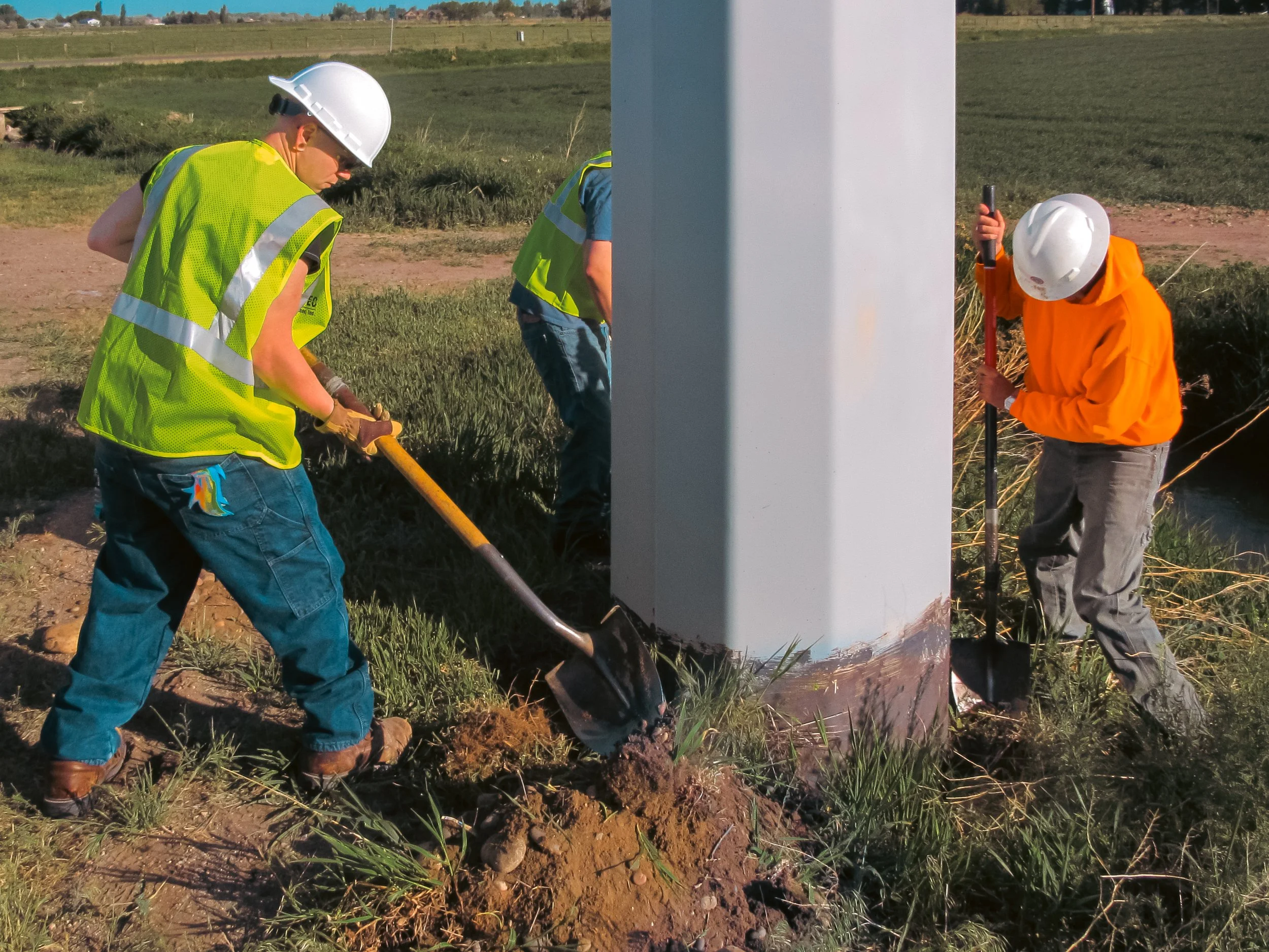 Three construction workers wearing helmets and safety vests working on the base of a tall metal structure in a field.