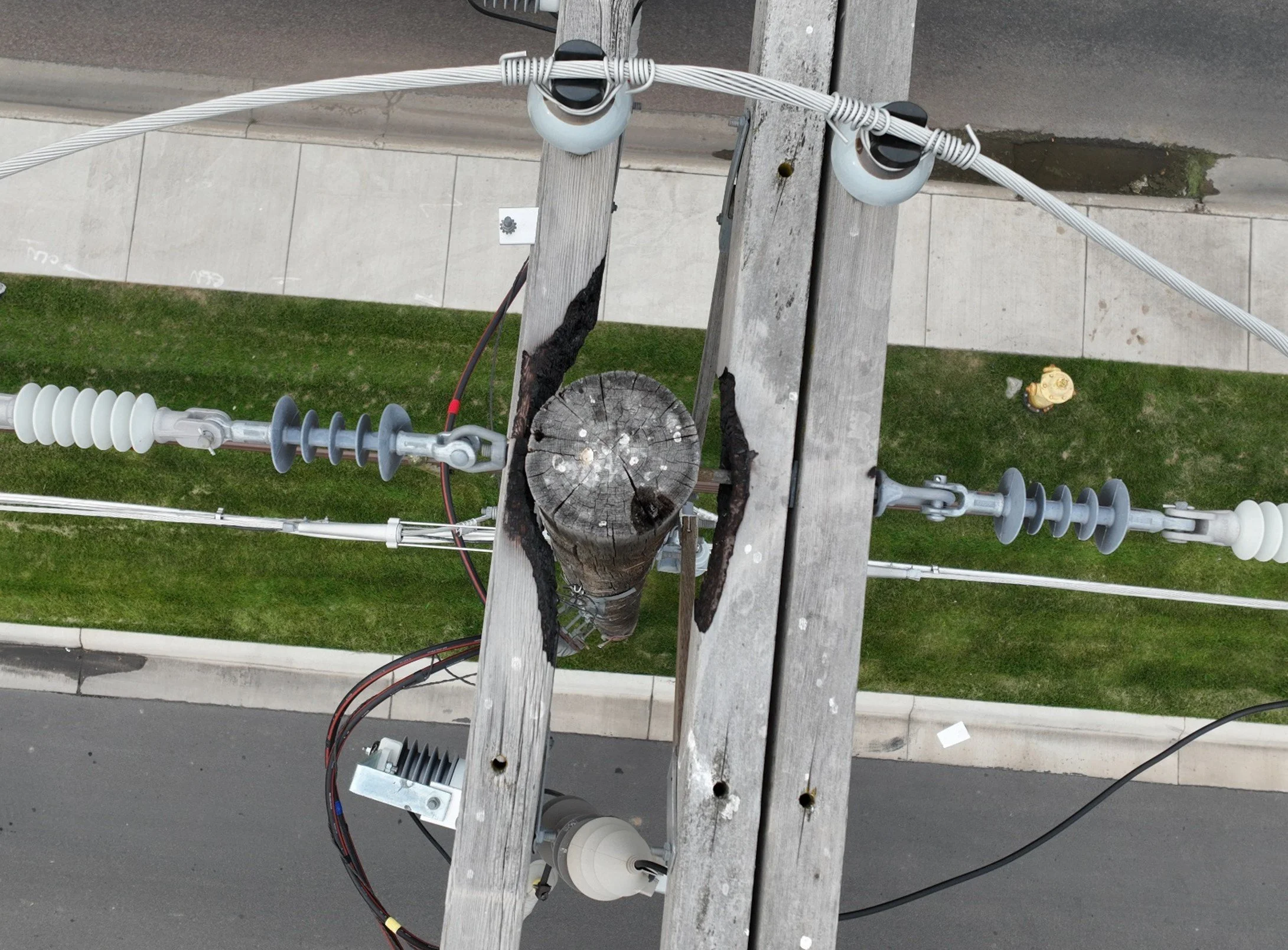 A photo taken from a utility pole showing electrical insulators, wiring, and a burned wooden crossarm, with a sidewalk, grass, and street below.