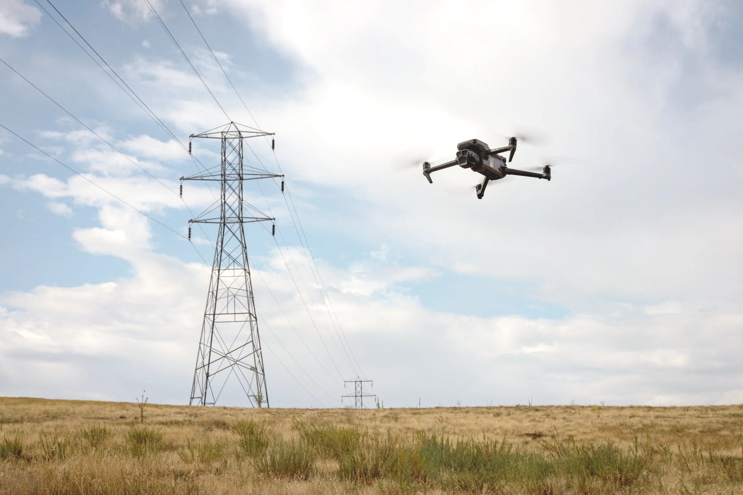 A drone flying near a high voltage power line tower in an open field under a cloudy sky.