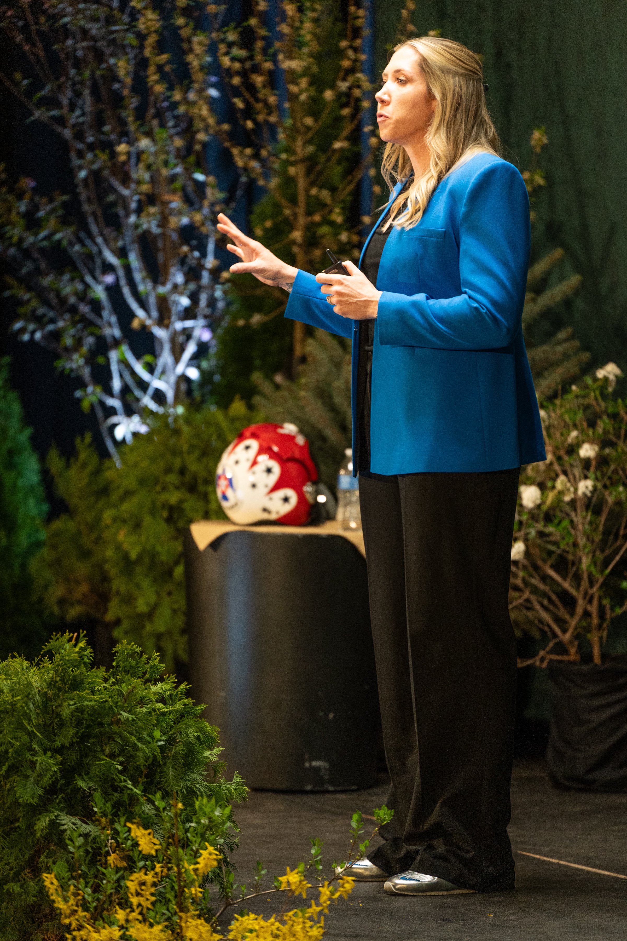 Woman giving a presentation on stage, wearing a blue blazer and black pants, with yellow flowers and greenery in the foreground and background.