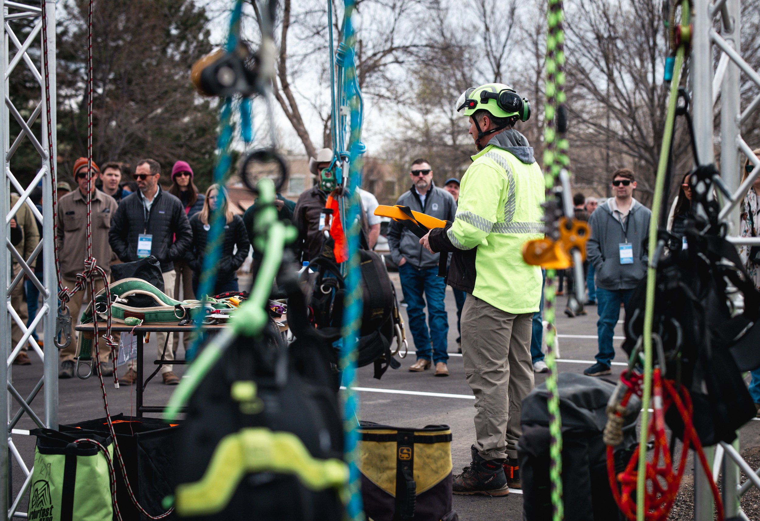 A man in a neon yellow safety jacket and helmet holds an orange tablet, surrounded by climbing gear and equipment at an outdoor event with a crowd of people in the background.
