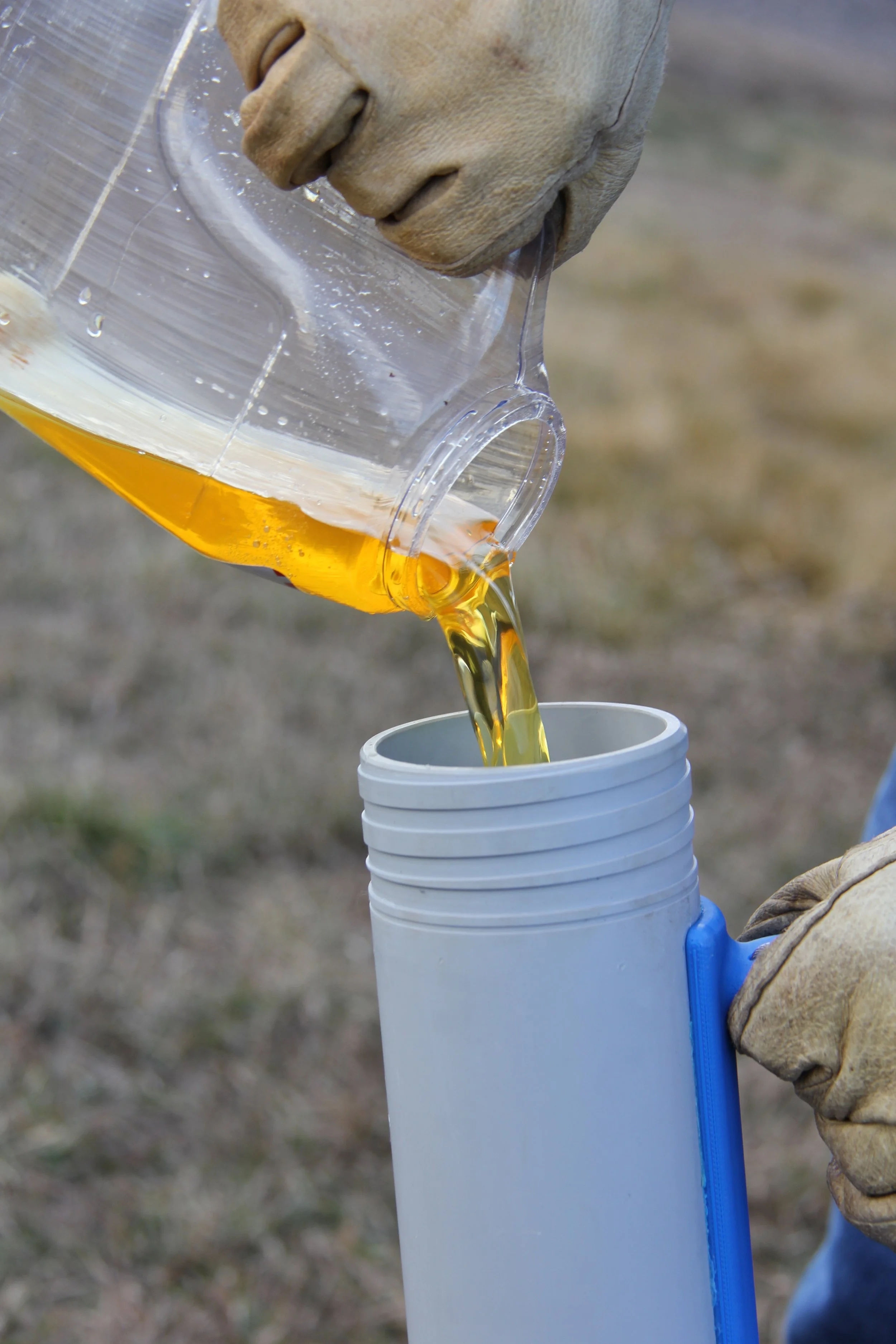 A person wearing gloves pouring beer from a plastic pitcher into a white thermos bottle outdoors.