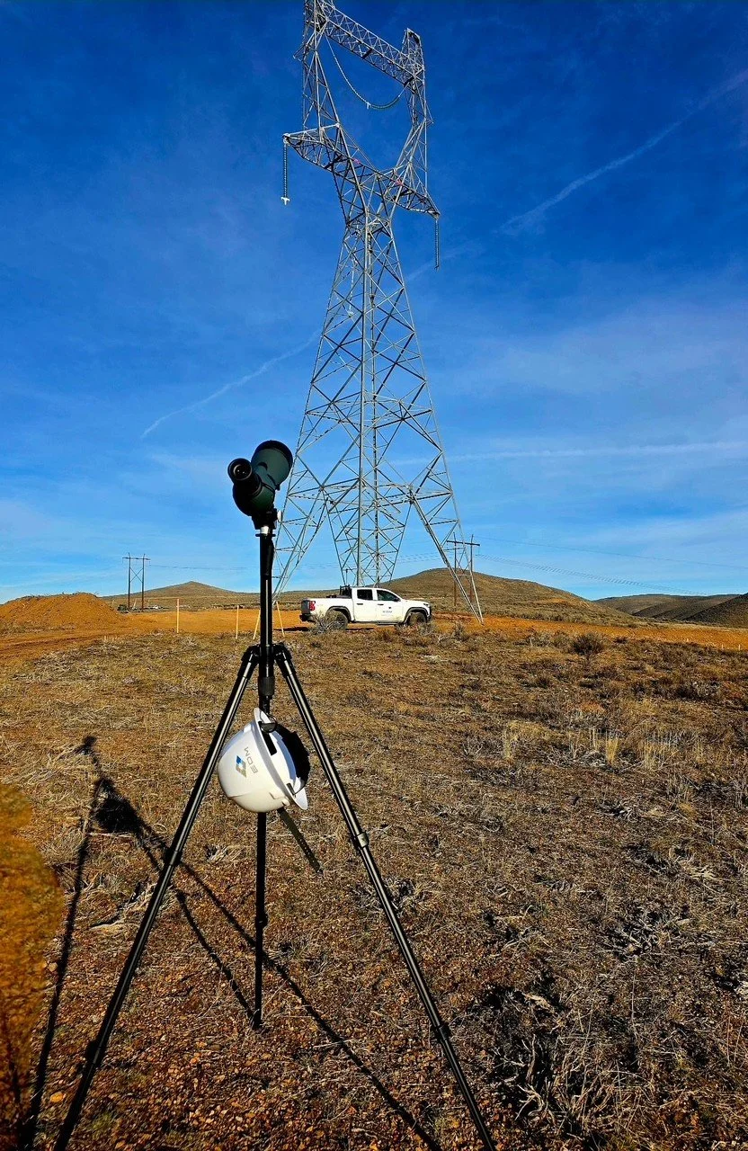 A wide landscape with a large electrical transmission tower, a white pickup truck nearby, and a telescope on a tripod in the foreground under a blue sky with some wispy clouds.