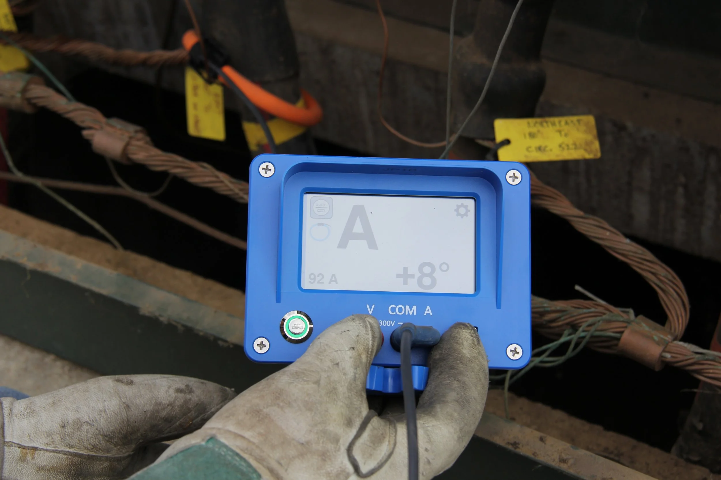 A person wearing dirty work gloves holding a blue multimeter with a digital display showing voltage measurements, in front of electrical wires and yellow tags attached to exposed wiring.