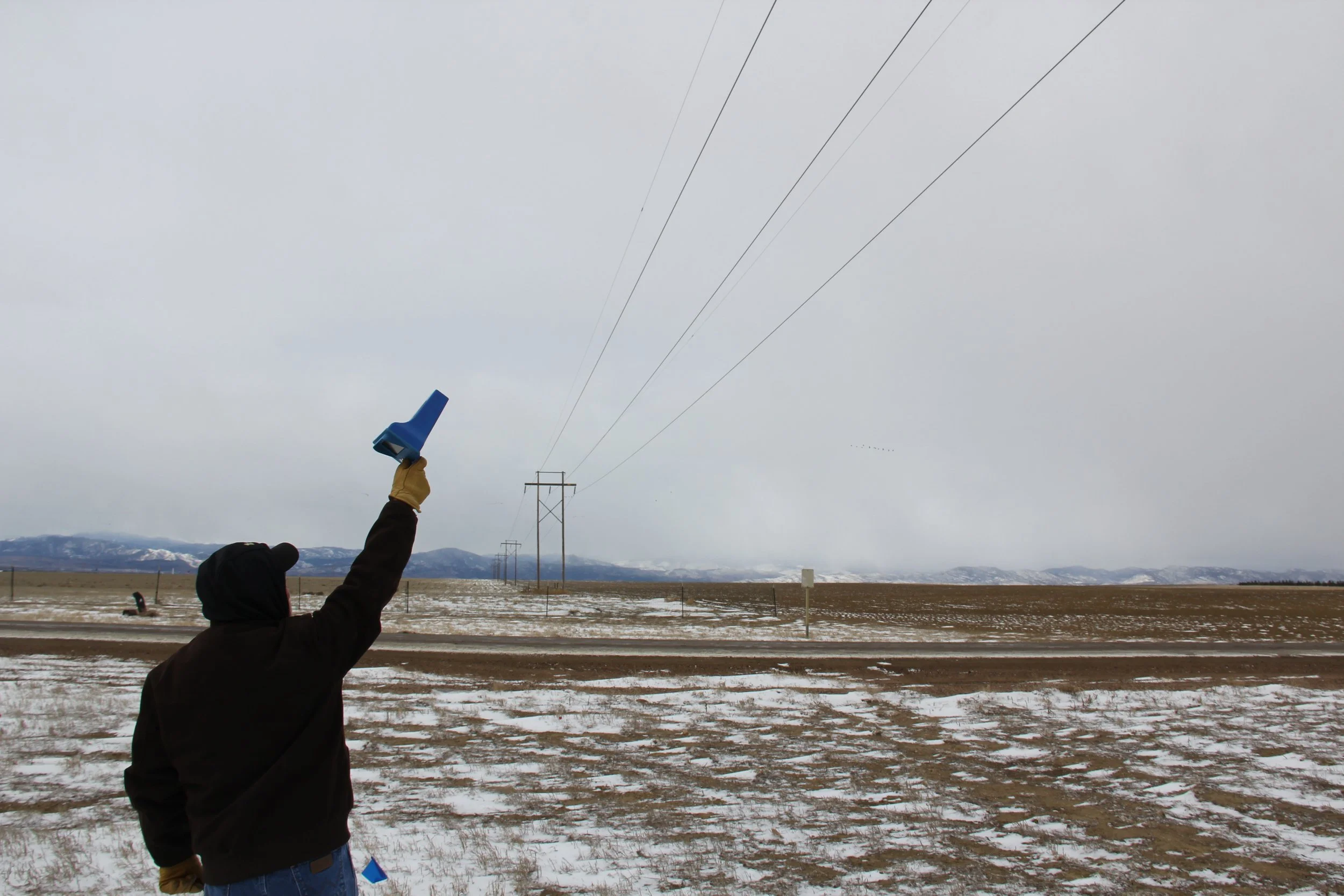 A person holding a blue and black radio transmitter with their hand raised in a cold, open field with snow, power lines, and mountains in the background.