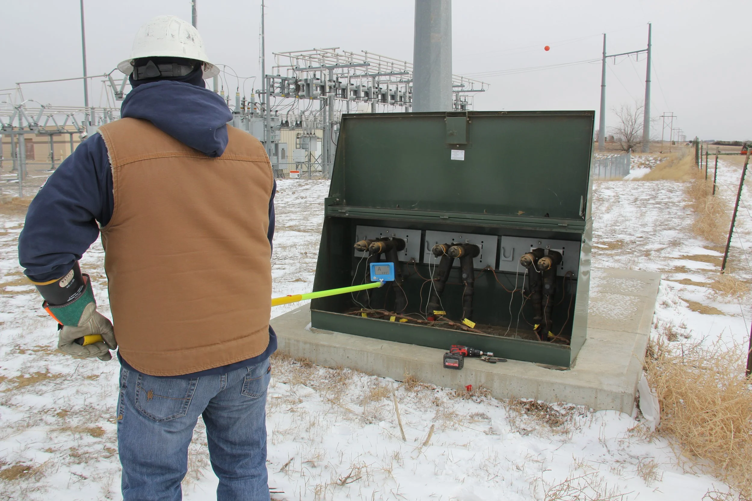 A worker wearing a white safety helmet, brown vest, and jeans working on electrical equipment outside in snowy weather, with electrical infrastructure in the background.