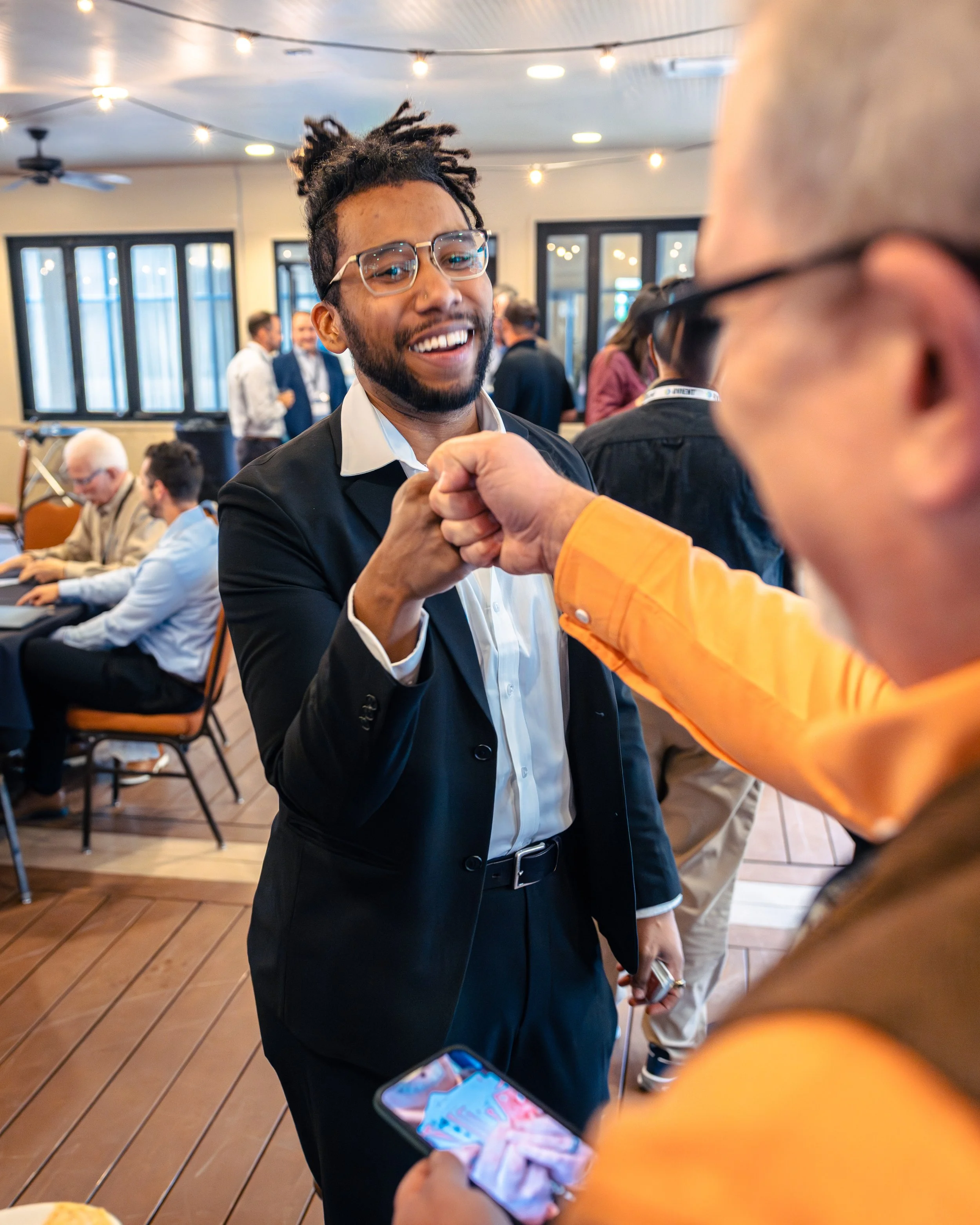 Two men are fist bumping at a gathering or celebration. The man in focus has dreadlocks, glasses, a beard, and is smiling. The other person's face is not visible, but they are wearing glasses and an orange shirt. In the background, people are sociali