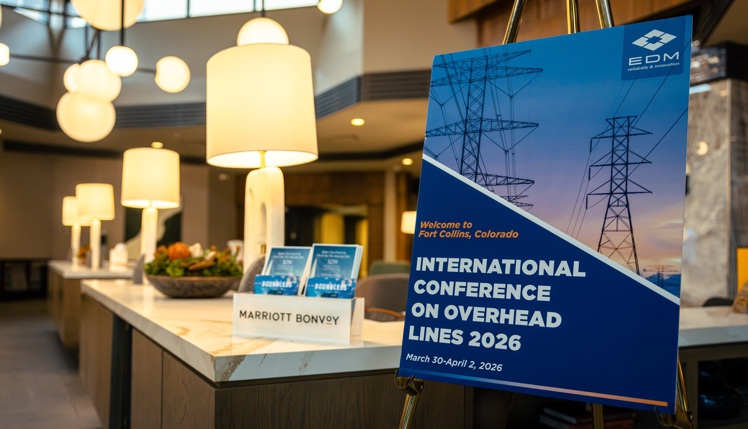 Conference registration desk at a hotel with a blue sign that reads 'International Conference on Overhead Lines 2026, March 30-April 2, 2026,' featuring an image of power poles. The desk has brochures and a flower arrangement, with hanging round lamps in the background.
