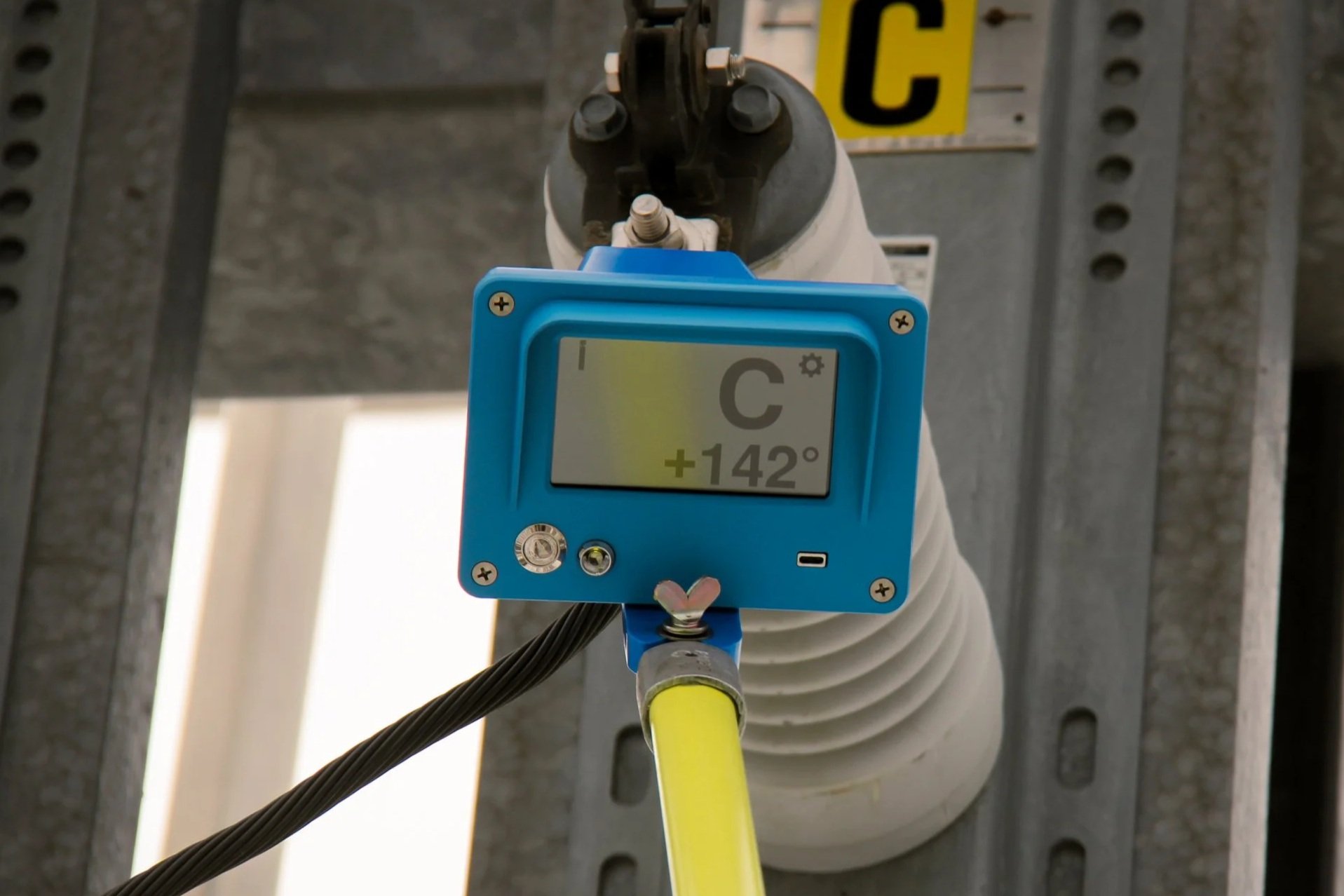 Close-up of an industrial temperature sensor or measuring device mounted on a pipe, showing a display reading 142 degrees Celsius, with wiring and mounting hardware visible, set against a background of an industrial metal framework.