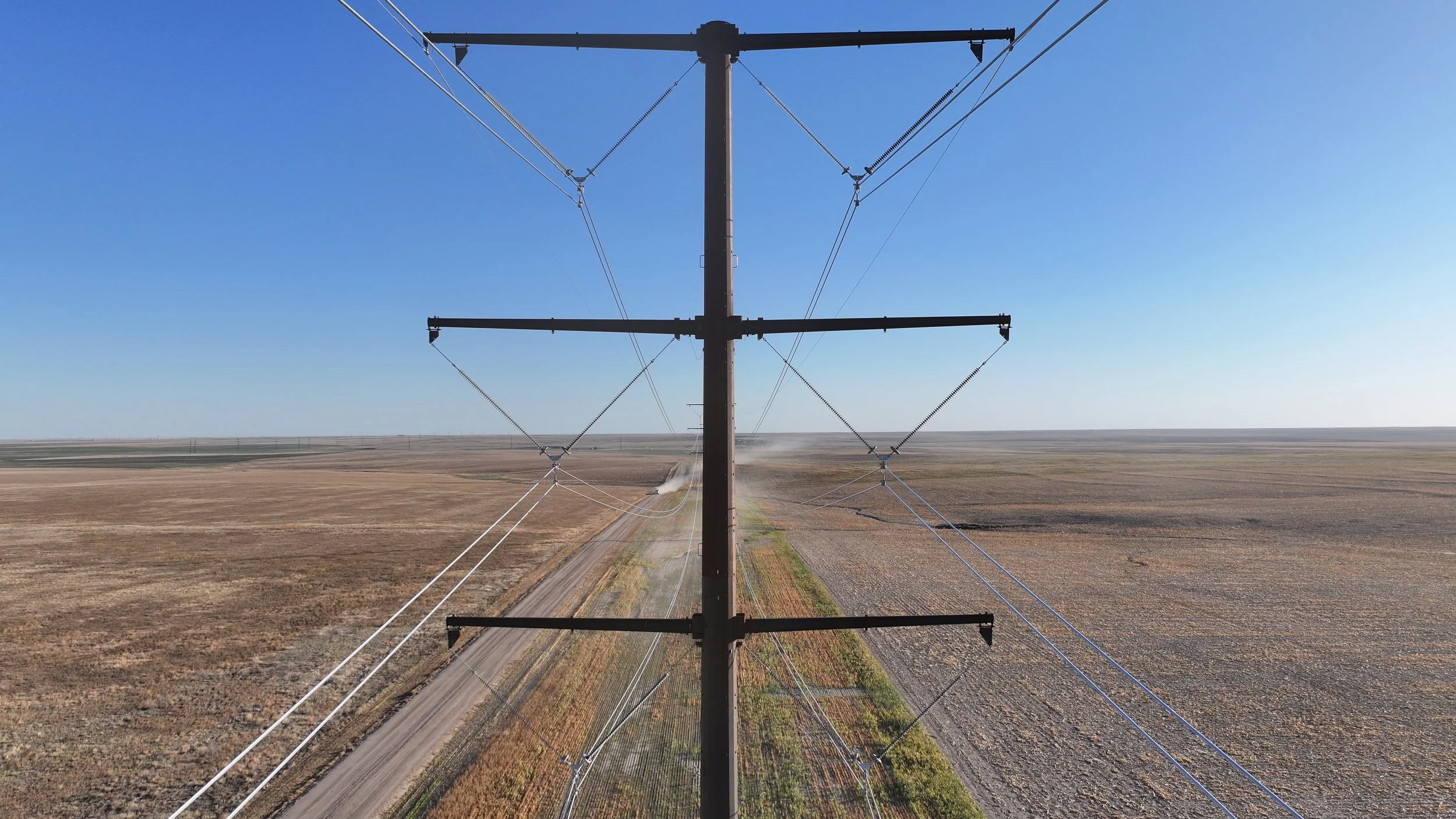 Electric power line pole in a rural field with a dirt road beneath, stretching toward the horizon on a clear day.