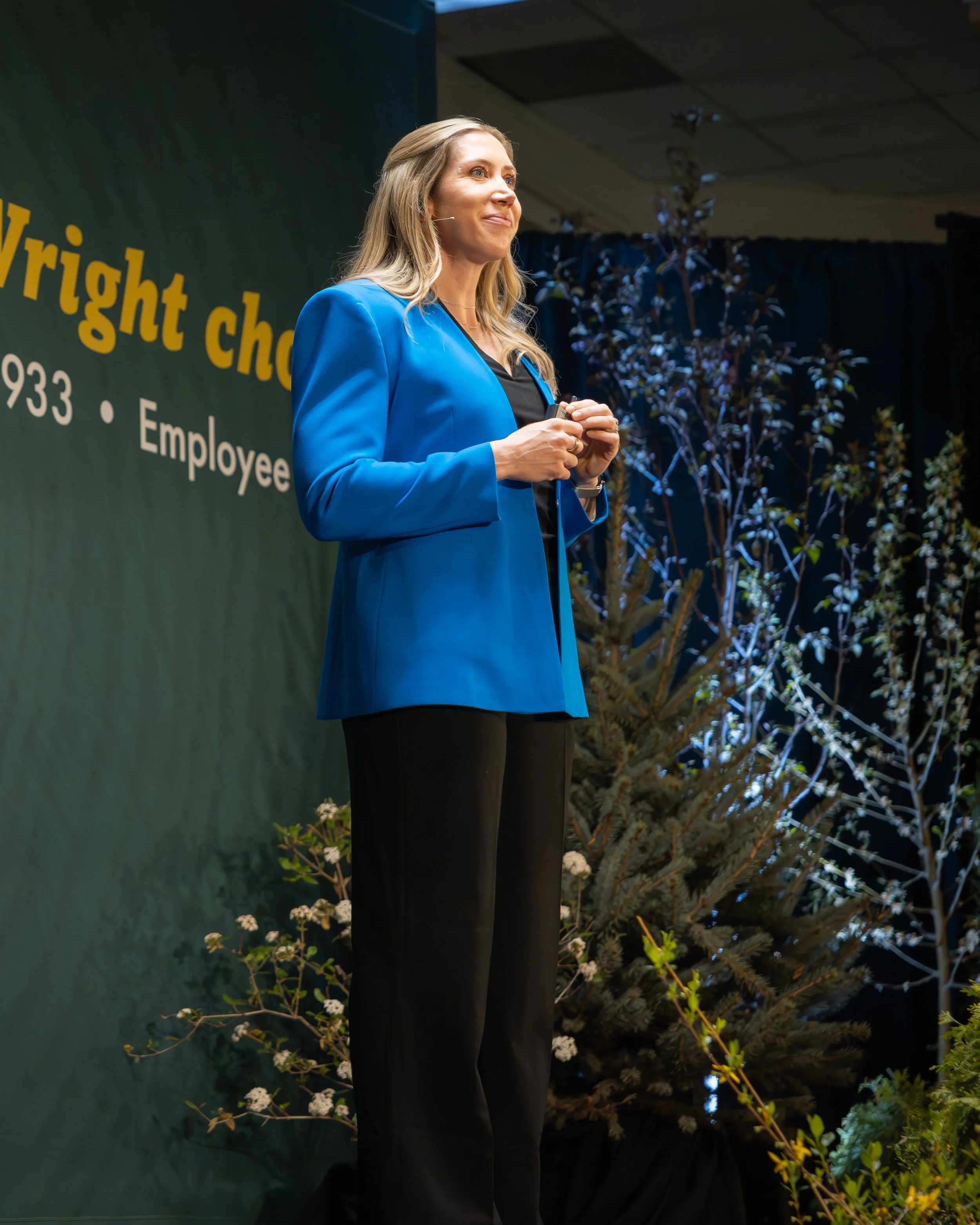 A woman in a blue blazer standing on stage during a presentation, with a dark background, plants, and flowers surrounding her.
