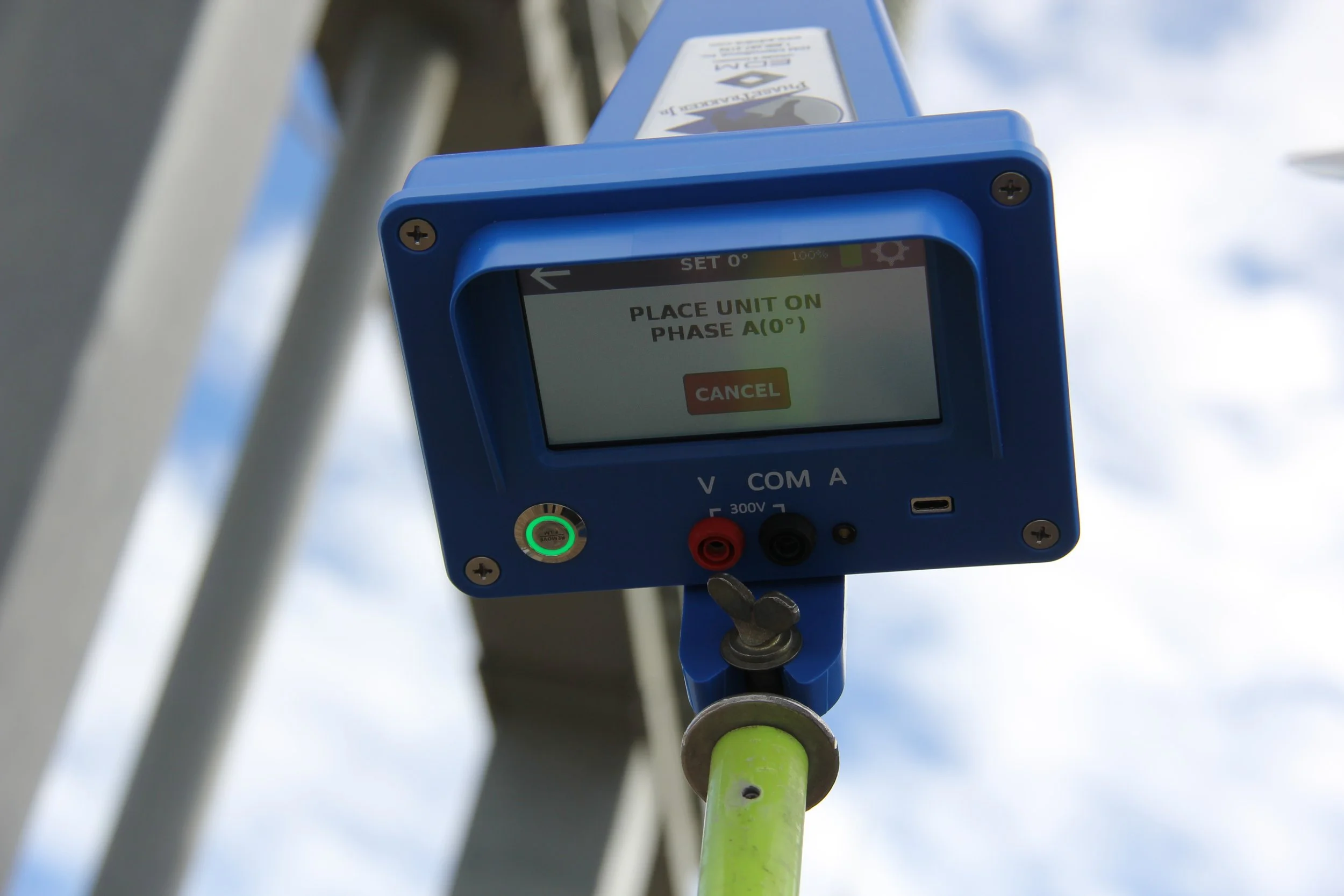 Electrical testing device mounted outdoors with a display screen showing instructions to place a unit on phase A 0°, with a cancel button below.