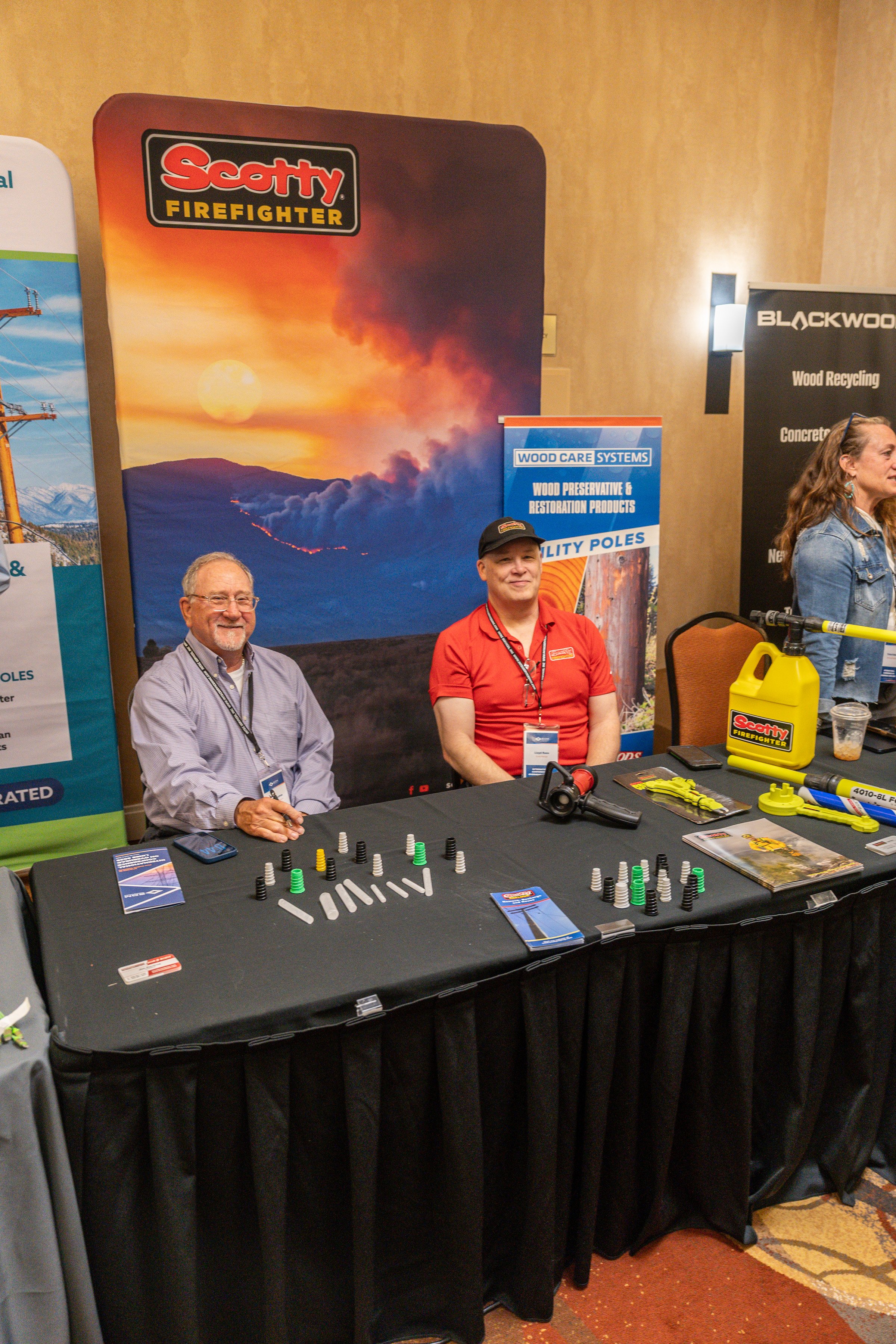 At an indoor exhibit booth, two men are seated behind a table displaying game pieces. The man on the left wears glasses and a striped shirt, and the man on the right wears a red shirt and cap. Behind them, colorful banners advertise firefighting and 