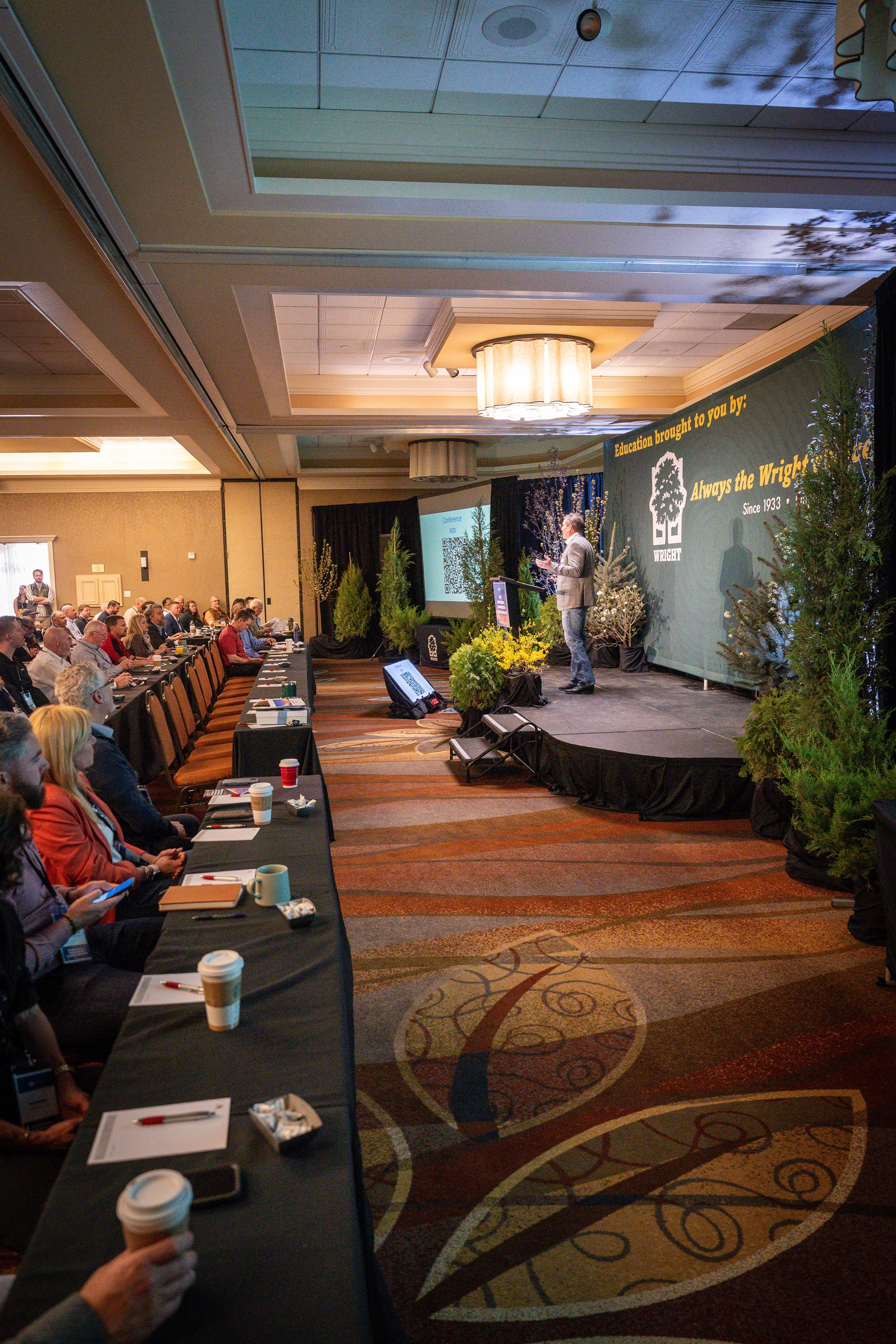 A conference room filled with attendees seated at long tables, facing a stage where a man is speaking at a podium. The stage has a banner with the logo and words 'Always the Wright' and decorative plants. The room has a patterned carpet and a drop ce