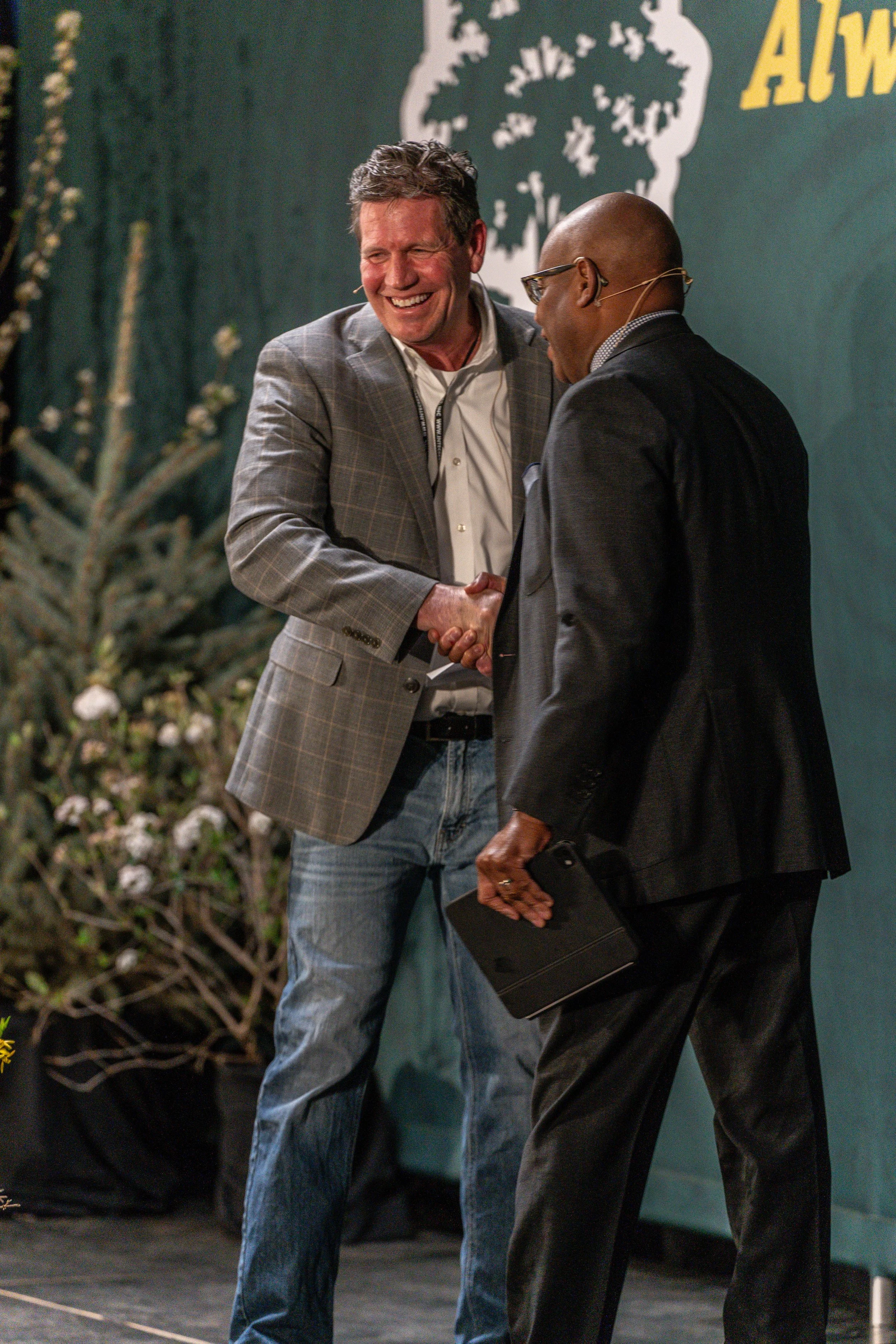 Two men shaking hands and smiling at an event. The man on the left has gray hair, a plaid suit jacket, and jeans. The man on the right is bald, wears glasses, a dark suit, and holds a black folder. A backdrop with a tree logo and yellow text is visib