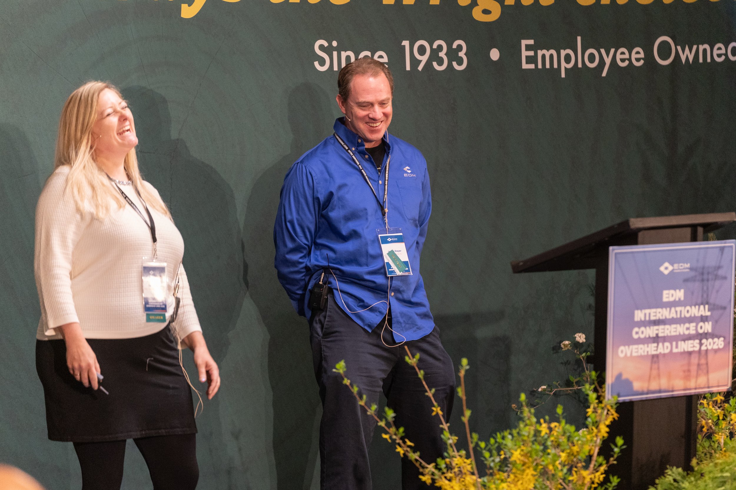 Two people, a woman and a man, standing on a stage at a conference. They are smiling and appear happy. The woman has long blonde hair and is wearing a cream-colored sweater with a black skirt. The man has short brown hair and is wearing a blue shirt 