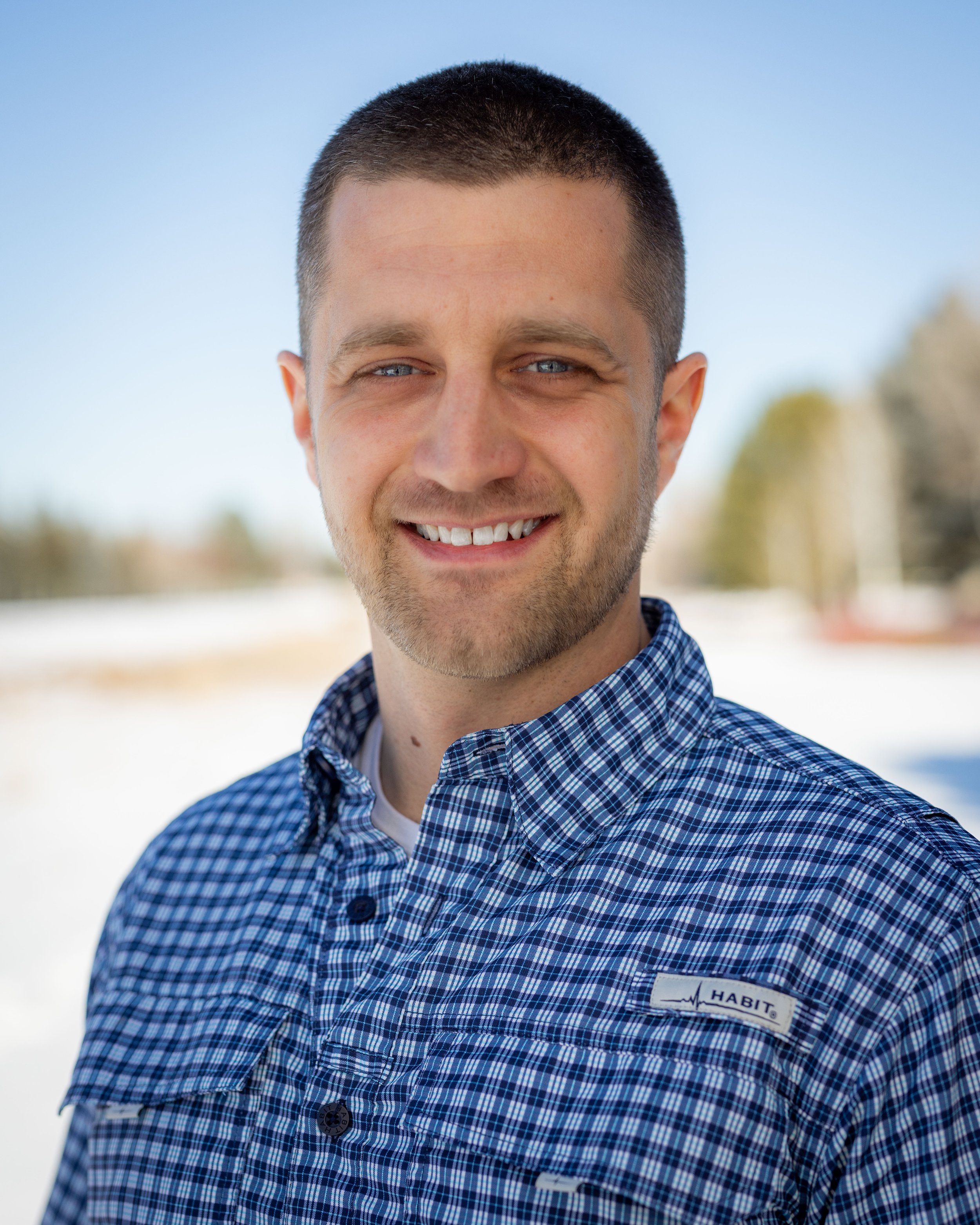 A young man with short brown hair, blue eyes, and a beard, smiling outdoors on a sunny day, wearing a blue checkered shirt, with a blurred background of trees and sky.