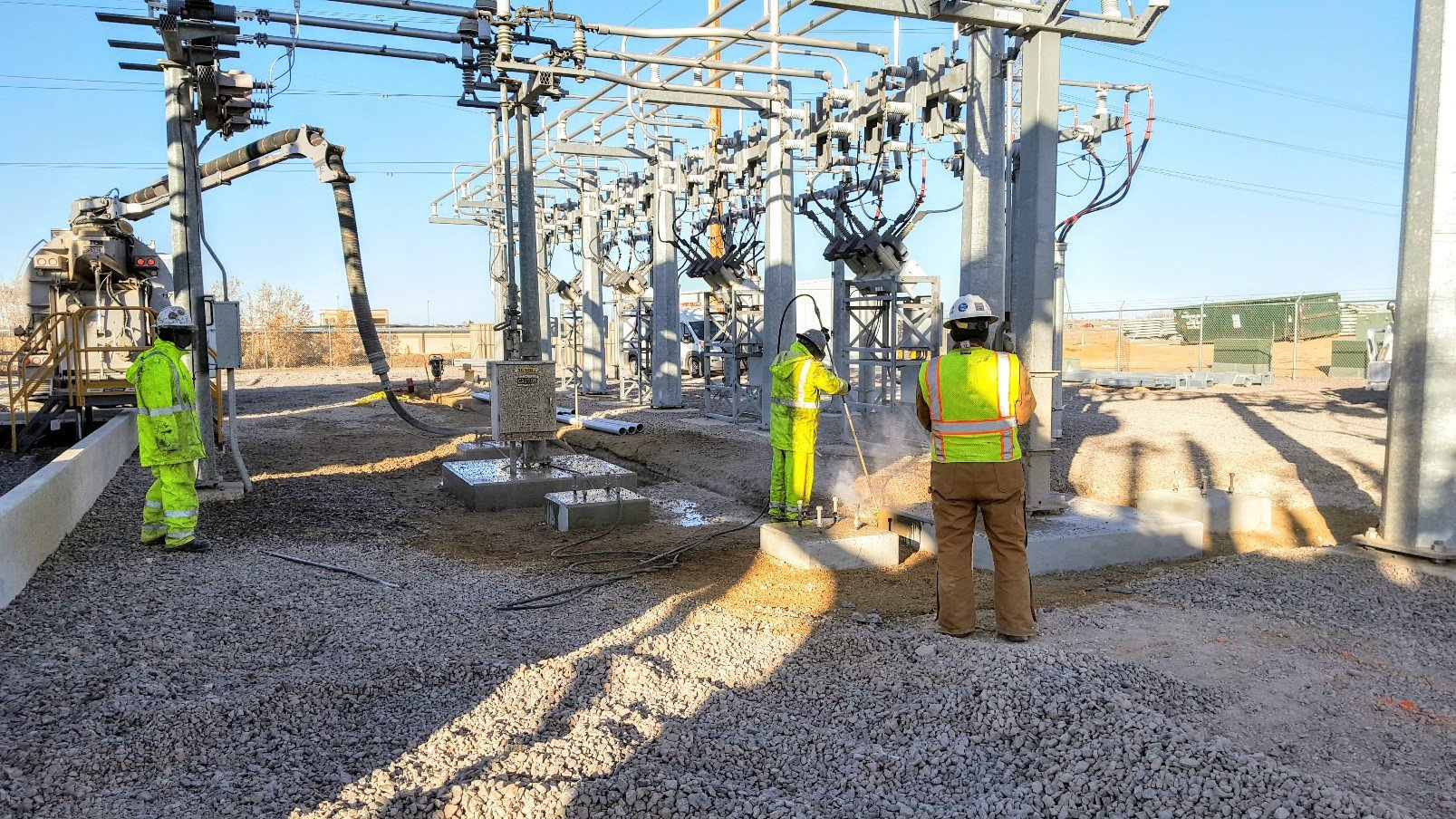 Construction workers in safety gear working on electrical substation with transformers and power lines.