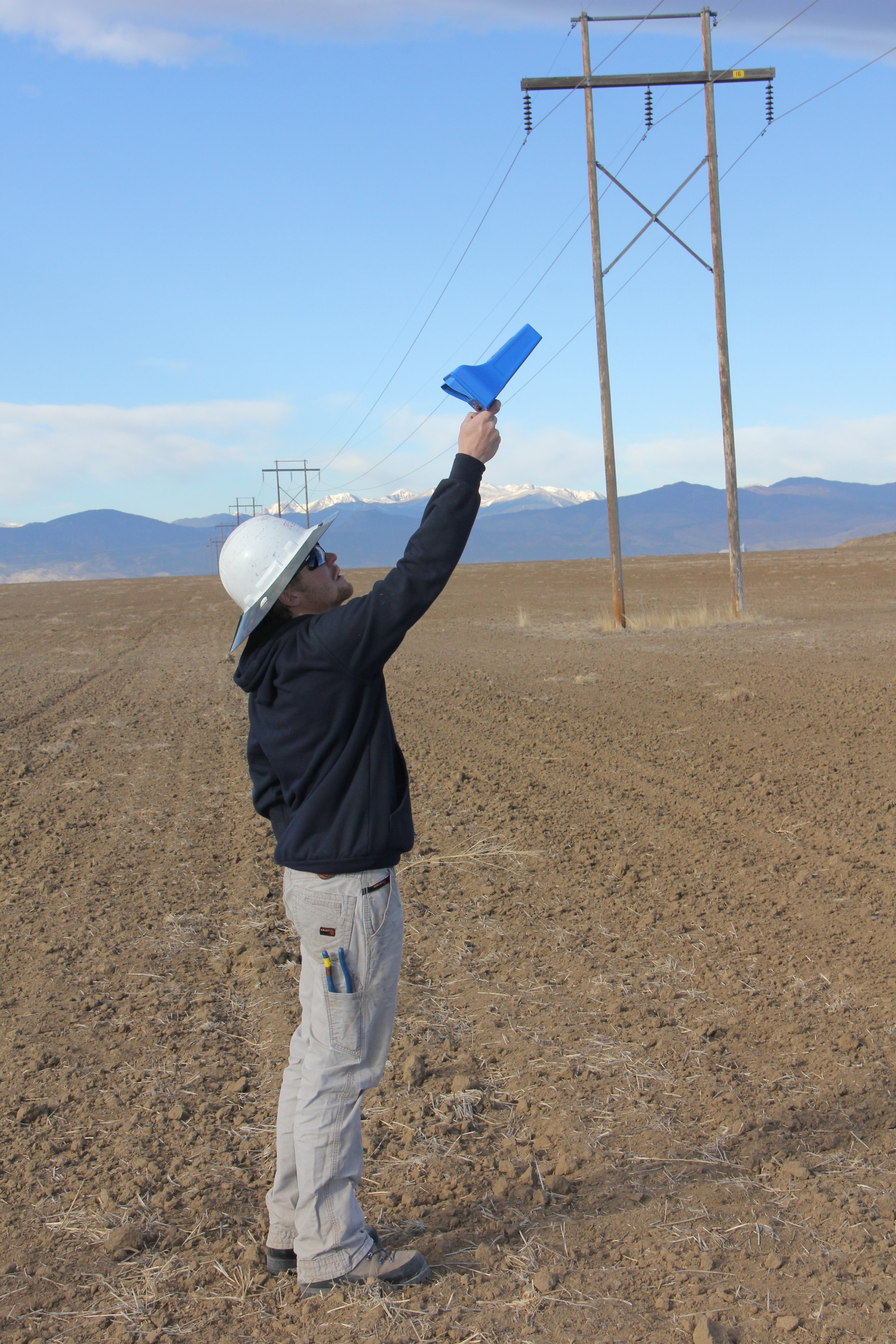 A man wearing a white hard hat, sunglasses, and work clothes stands in a field, holding a blue flag in his right hand, extended upward towards electrical poles and wires across an open landscape with mountains in the distance.
