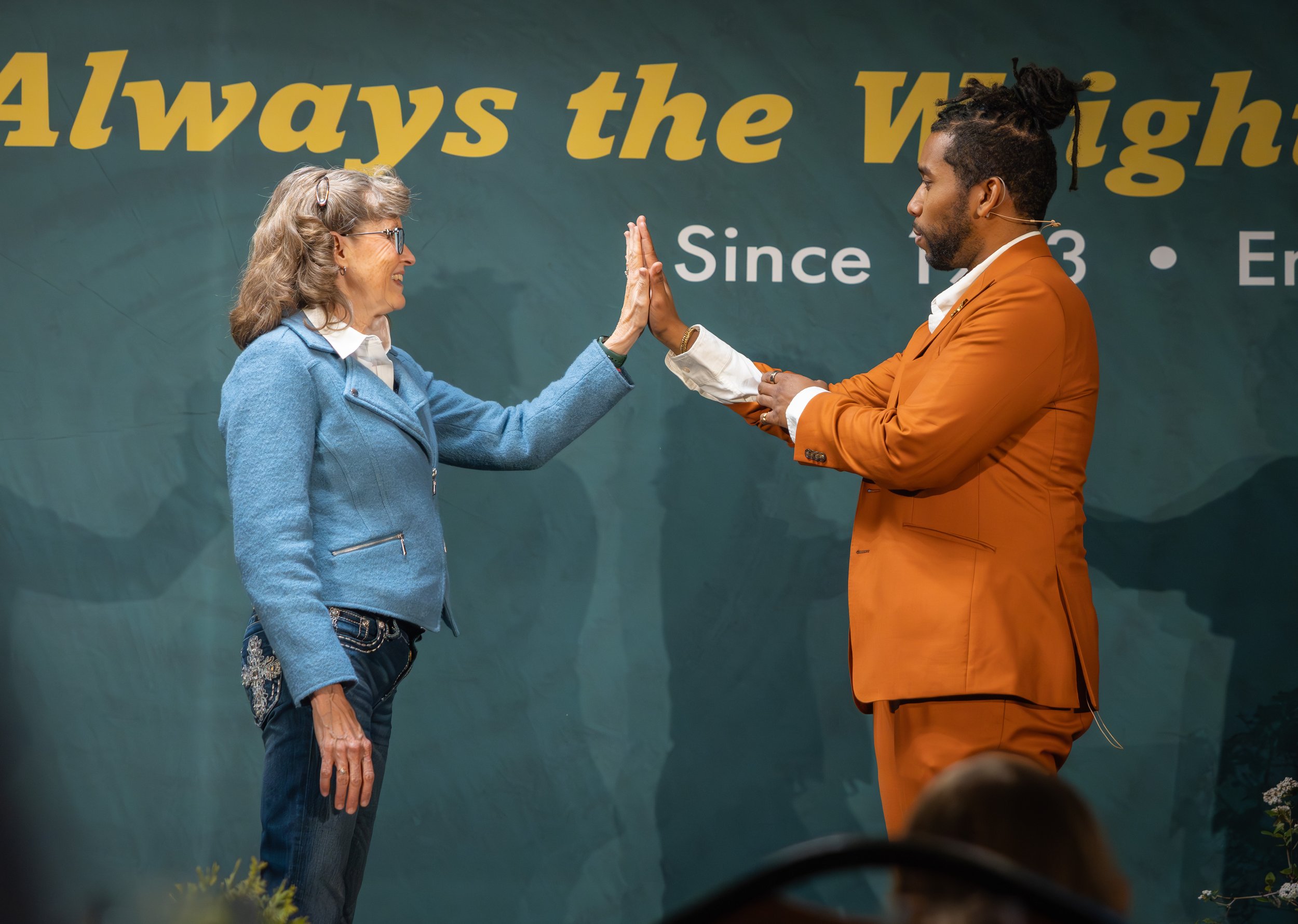 An older woman and a man with dreadlocks high-five each other on stage in front of a dark background with yellow text that reads 'Always the Wigh...'. The woman is wearing glasses, a blue jacket, and jeans. The man is dressed in an orange suit with a