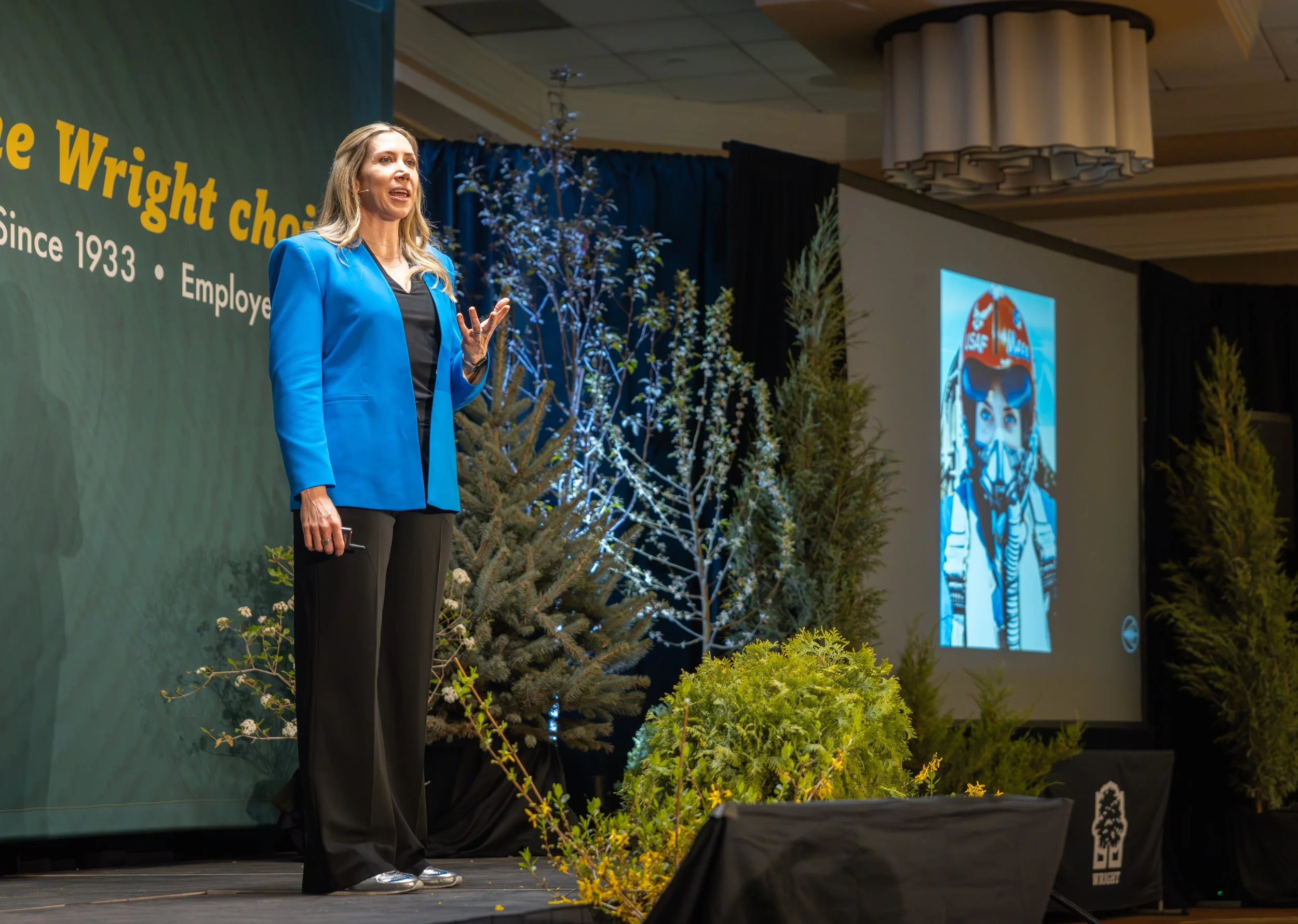 A woman in a blue blazer giving a speech on stage decorated with plants and trees, with a large screen displaying an image of a person in goggles to her right.