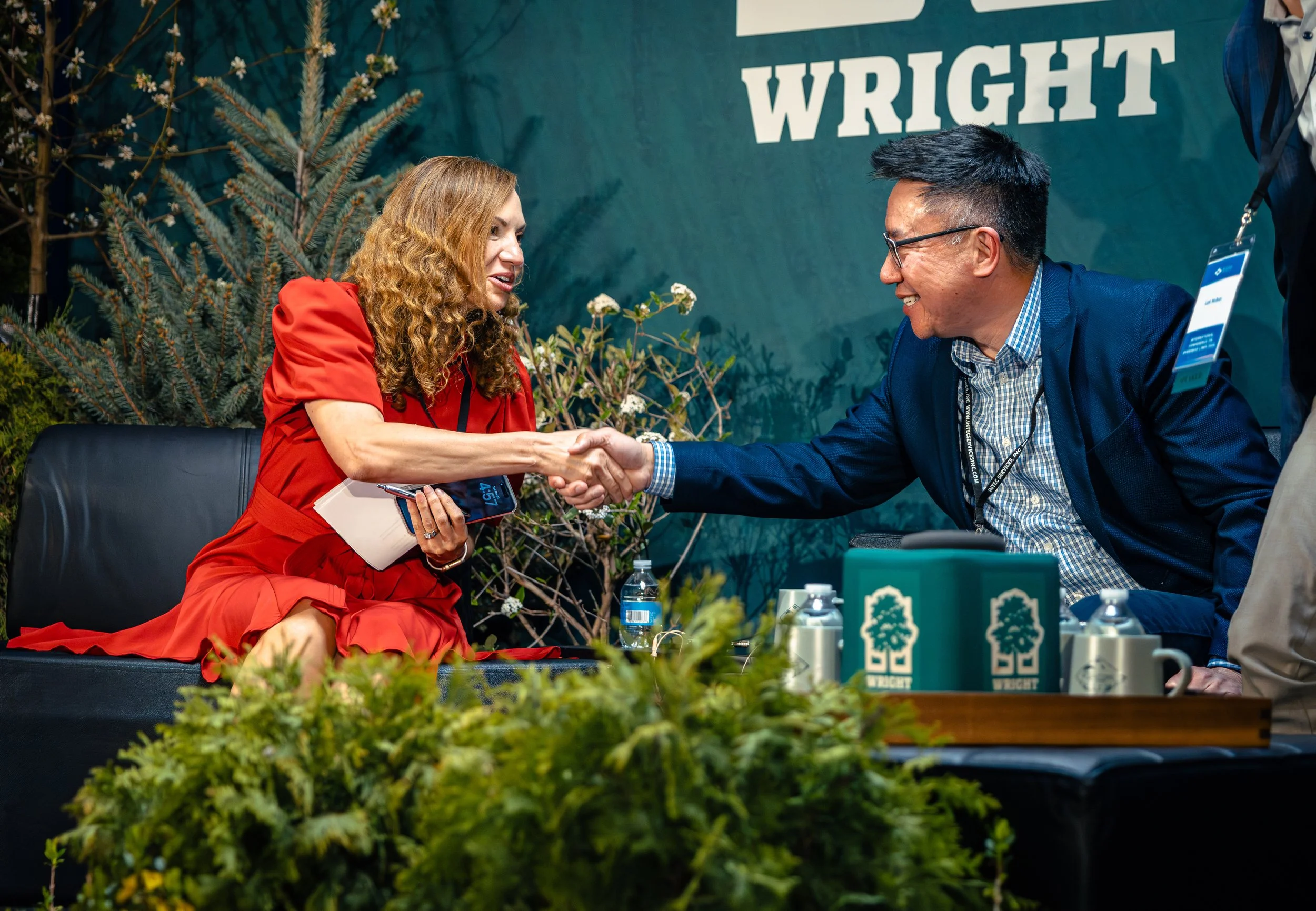 A woman in a red dress and a man in a blue jacket shaking hands at an event with a green backdrop and floral decorations.