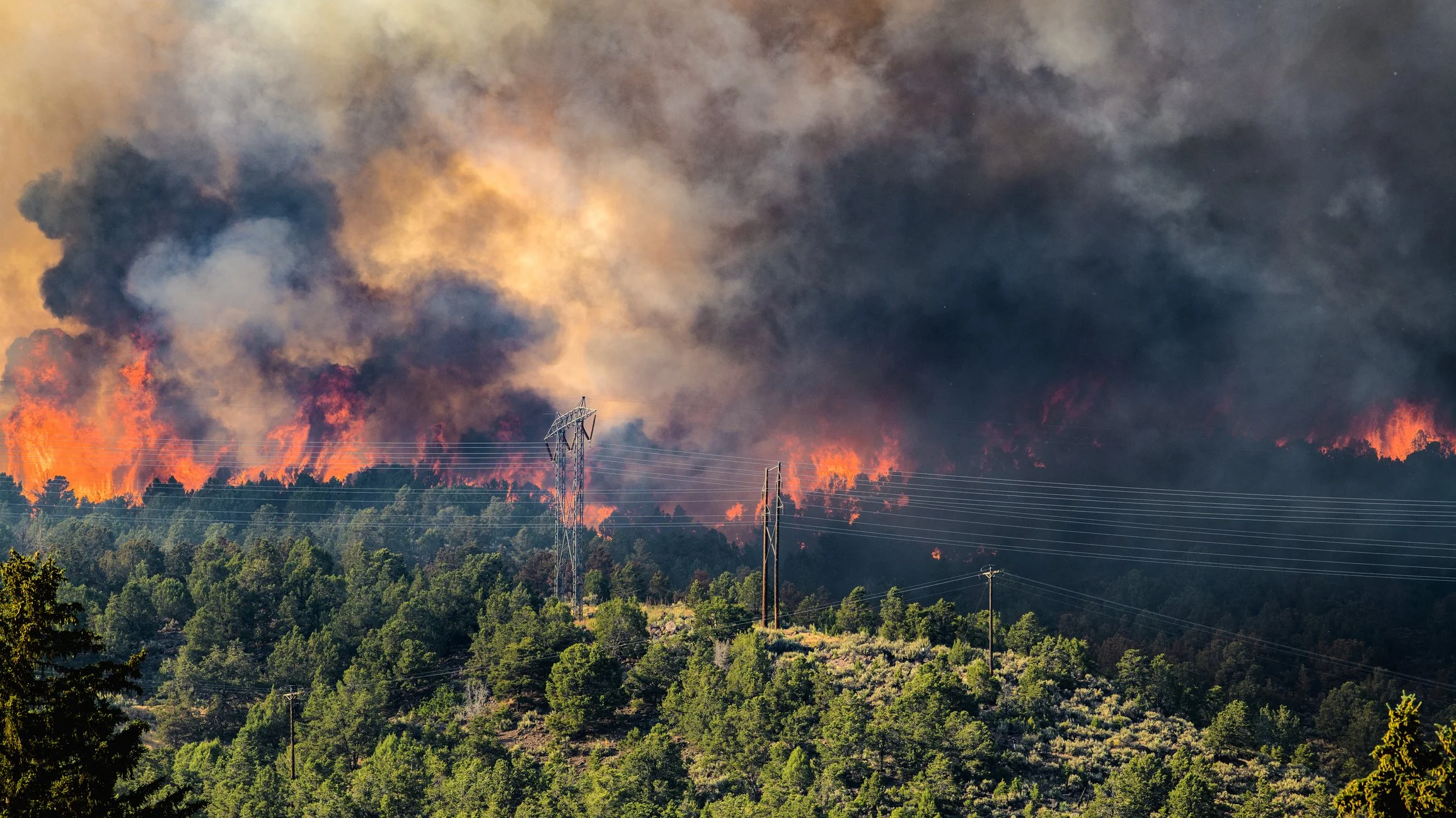 Wildfire burning on a forested hillside with thick black smoke and bright orange flames, and power lines and transmission towers in the foreground.
