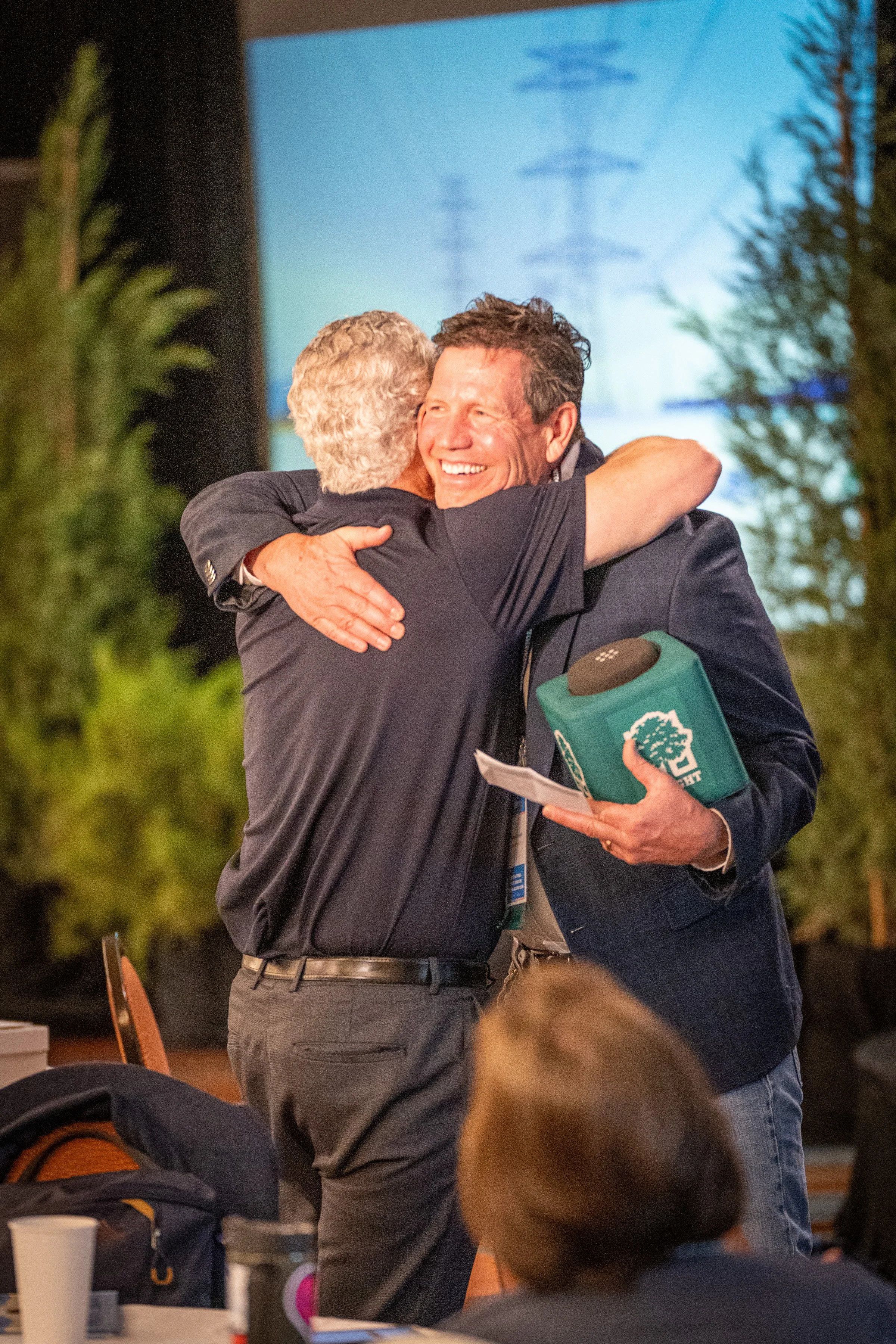 Two men are hugging and smiling at each other during a gathering or event. One man has gray hair and the other has dark hair. There are trees, a table with cups, and a large screen or projection in the background.