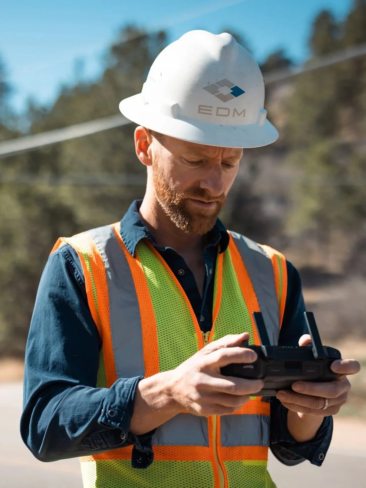 A man wearing a white safety helmet with the logo 'EDM' and a high-visibility orange and yellow vest over a blue shirt, operating a remote control device outdoors on a sunny day.