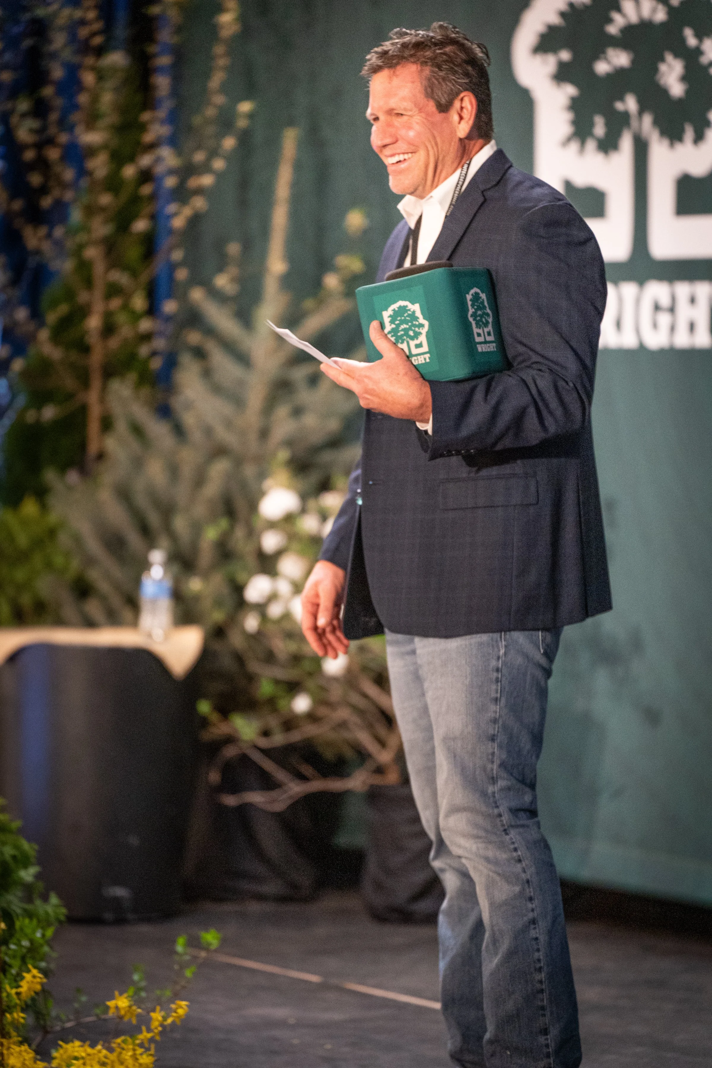 A man in a dark blazer and grey jeans holding a book or folder at an event. There is a logo with a tree and the words 'The Right' in the background, and some flowers and plants in the foreground.