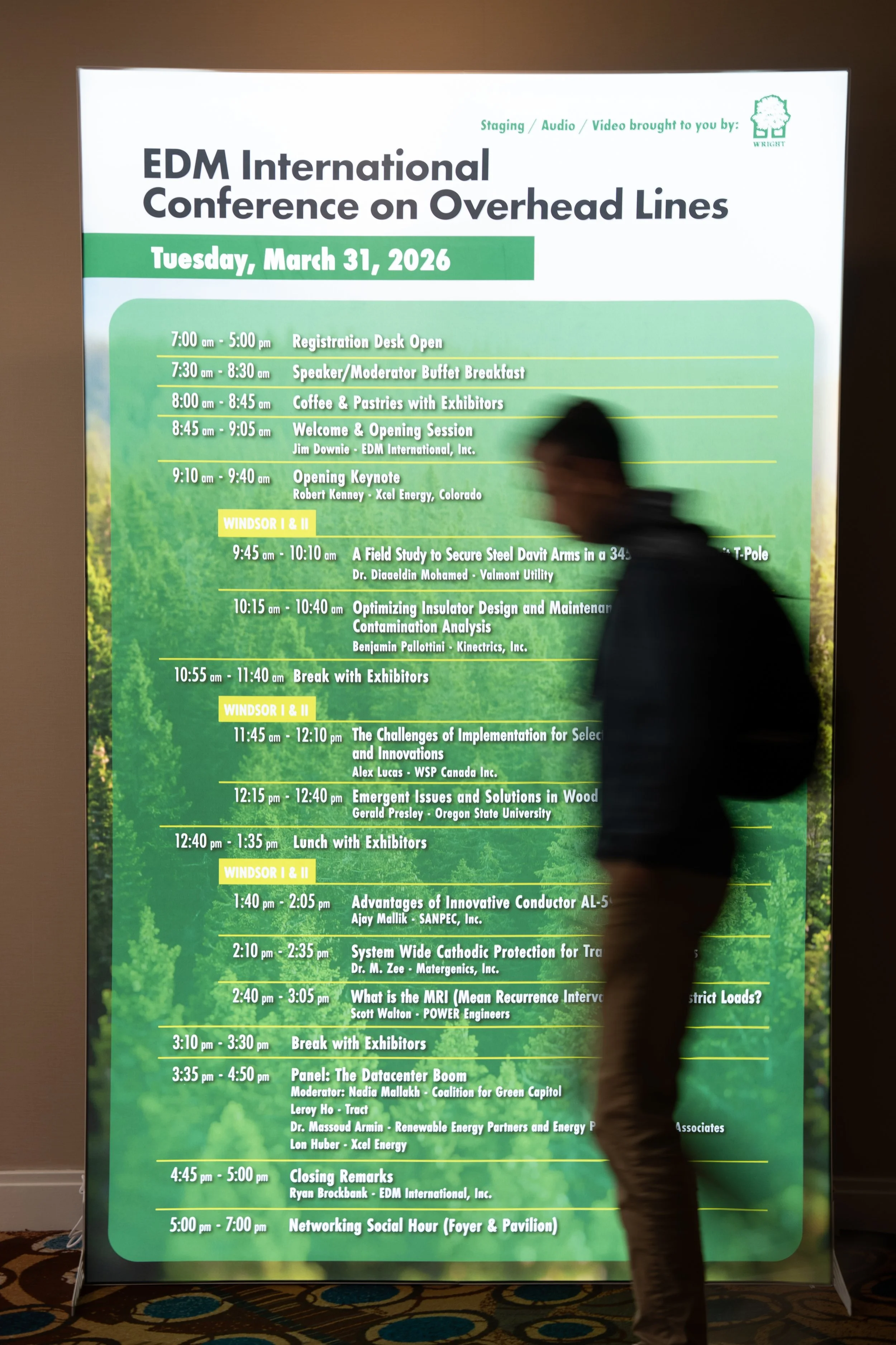 Large digital conference schedule board displaying the agenda for the EDM International Conference on Overhead Lines, scheduled for Tuesday, March 31, 2026, with session times, topics, and speakers. A blurred person walks past in front of the board.